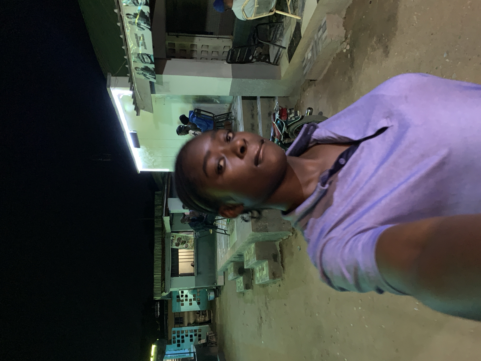 The image presents a selfie of a person with dark hair and skin, wearing a light purple top. In the background, there's an outdoor scene taken at night with artificial lighting. To the left, there is a building with several windows and what appear to be ventilation blocks on the bottom. Some people can be seen seated around the building. A motorcycle is also present. The environment appears to be a street or public space in the area of New Stadium, Nigeria.