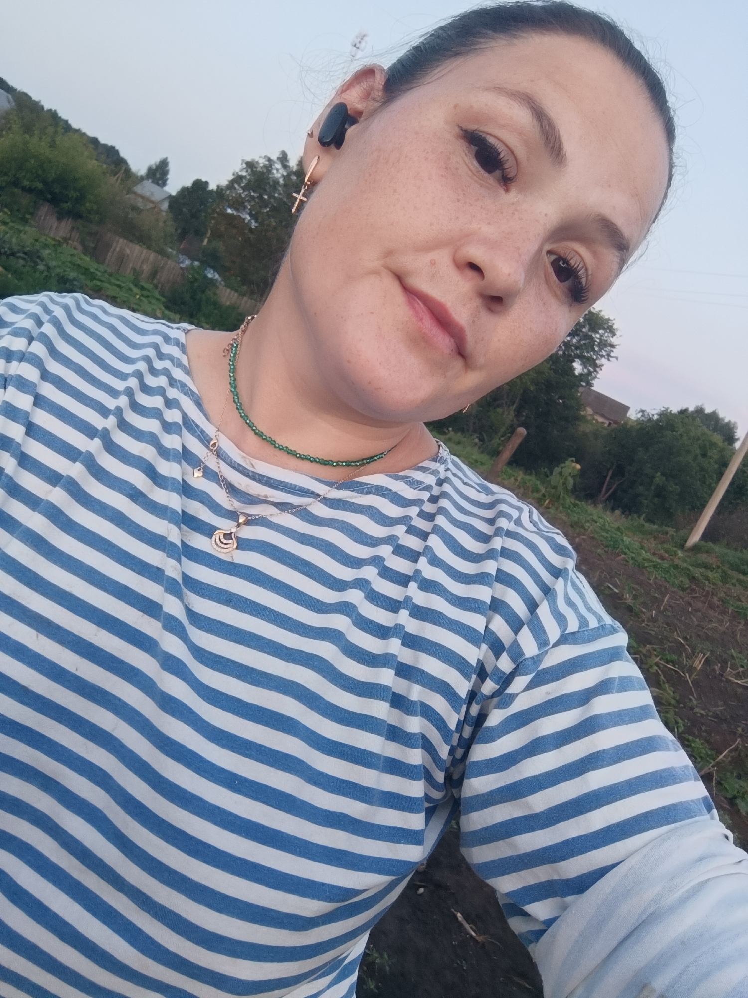 In a medium shot, a woman with her hair pulled back and freckles across her face is captured outdoors, with a slightly blurred background suggesting a rural or suburban setting. She is wearing a blue and white striped t-shirt and two necklaces, one a delicate gold chain with a pendant and the other a strand of green beads. She also has a small cross earring in her right ear and a black earbud. The background shows a gentle slope with trees and some structures, hinting at the outskirts of a town. The lighting suggests it is either early morning or late evening. The overall tone of the image is casual and candid.