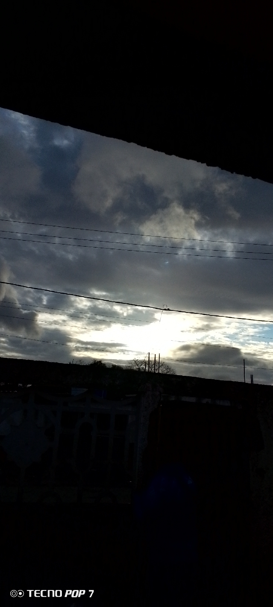 The image shows a cloudy sky with dark clouds in the foreground and brighter, white clouds closer to the horizon. Several utility lines run horizontally across the scene. Below the sky, there is a silhouette of buildings and trees. The foreground is dark, partially obscuring details. The "TECNO POP 7" watermark is visible in the bottom left corner. The location is Altagracia, Venezuela.