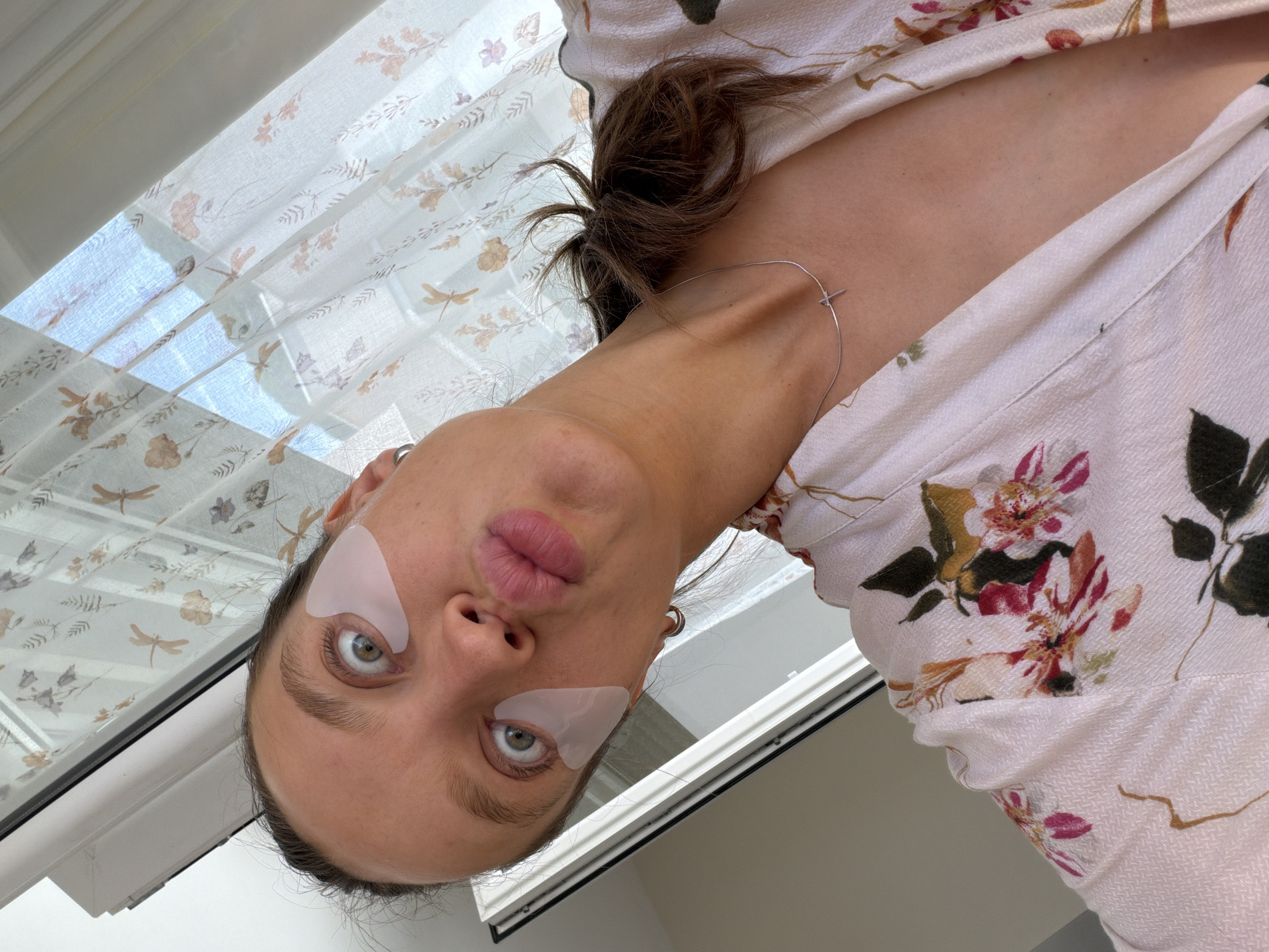The image is a close-up, low-angle shot of a young woman, rotated 90 degrees clockwise, looking directly into the camera. She has wide, striking green eyes and her lips are slightly pursed, conveying a playful or surprised expression. Light pink or white under-eye patches are visible beneath both eyes, indicating a skincare routine. Her dark brown hair is pulled back, with a messy bun visible on her neck/shoulder. She wears a thin silver necklace with a small cross-shaped pendant and a hoop earring. Her garment is a light-colored (possibly off-white or cream) fabric with a prominent floral pattern in shades of pink, red, green, and brown, suggesting a dress or top.
In the background, a window is visible, covered by a sheer, white curtain adorned with a delicate pattern of dragonflies and small floral motifs. Bright natural light filters through the curtain, creating a soft glow and indicating that it is daytime. A white window frame runs along the edge of the scene, confirming an indoor setting. The image captures a casual, intimate moment of self-care. This scene is set in Budva, Montenegro. There is no visible text in the image.