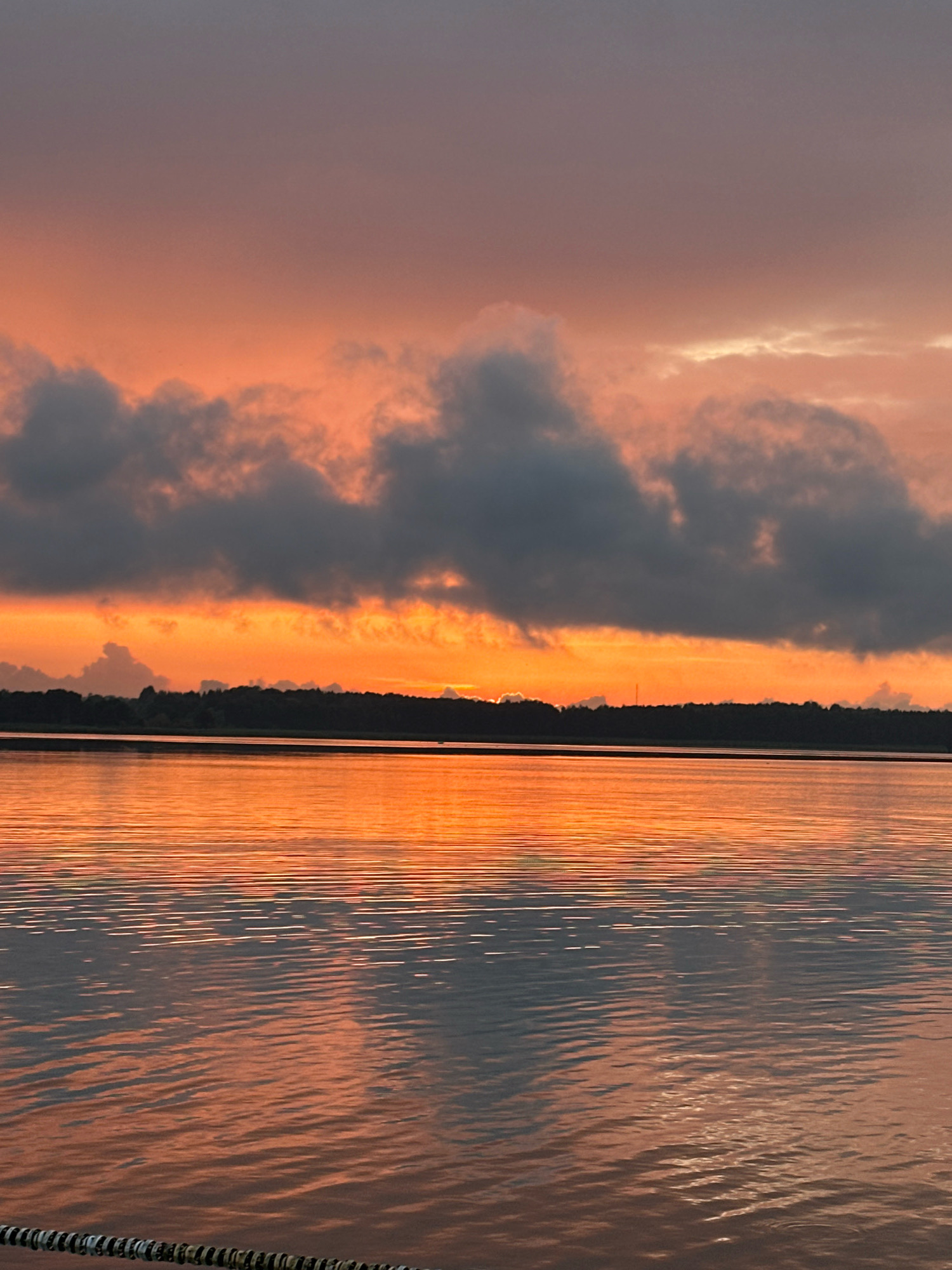 The image depicts a tranquil lake scene at sunset, with a treeline and dark clouds in the distance. The sky is ablaze with vibrant orange and pink hues, reflecting beautifully on the calm water. The foreground shows a partially submerged rope or chain, hinting at a dock or a mooring. The scene is serene and evokes a sense of peace and natural beauty.
The specific location in Tallinn, Estonia cannot be determined from the image alone, as it is a generic landscape scene that could be found in many places with lakes and sunsets.