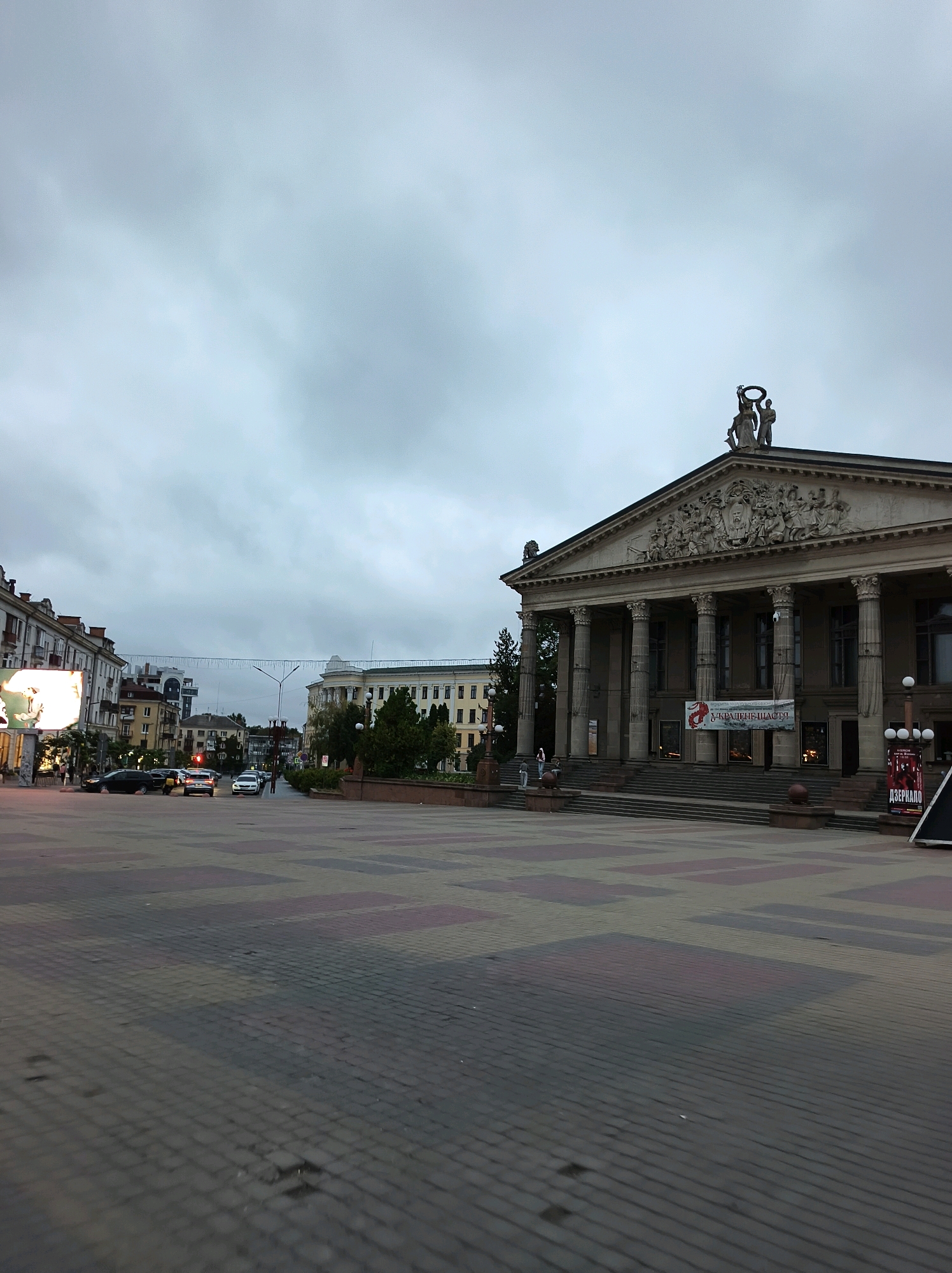 The image depicts a frontal view of the Ternopil Academic Drama Theatre, a grand building with a neoclassical facade featuring tall columns and intricate sculptural details. Two statues stand atop the pediment, one holding a laurel wreath. A banner with Cyrillic text, possibly advertising a performance, hangs in front of the entrance. The theatre is situated in a spacious, paved plaza, with cobblestones laid in a geometric pattern. To the left of the theatre, a street is visible with several cars, including a black sedan, a white car, and a yellow taxi. Buildings line the street, with one featuring a large illuminated billboard on the left. In the distance, more urban structures and trees are present under an overcast sky, suggesting it is either early morning or late evening. A few individuals are seen in the plaza, and on the steps of the theatre. The overall atmosphere is subdued, possibly due to the cloudy weather.