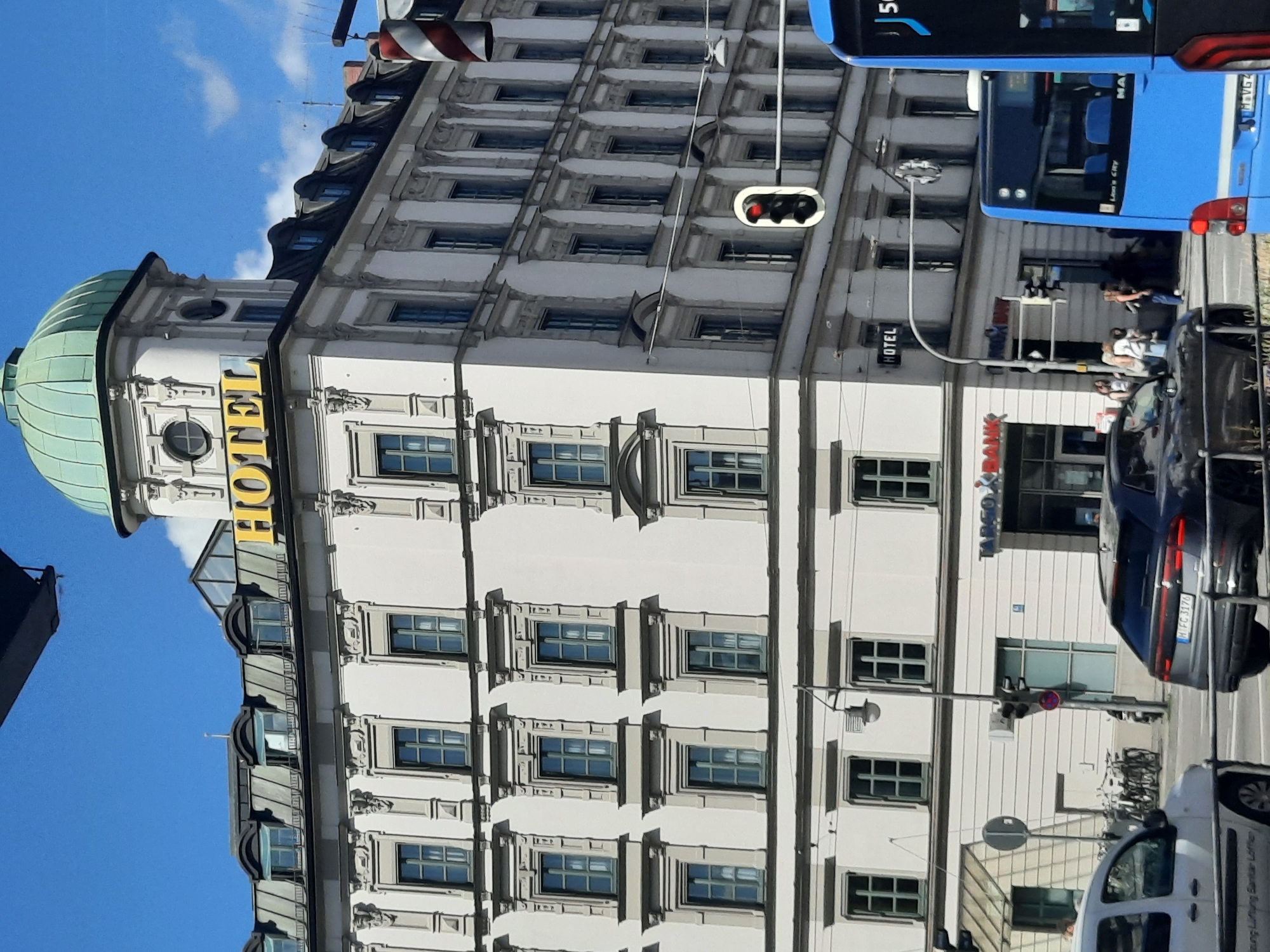A street view captures a bright, sunny day in an urban setting. The focus is on a large, ornate building with a light-colored facade, adorned with numerous windows and decorative elements. A prominent yellow sign with the word "HOTEL" is visible near the top of the building, below a green domed roof. In the foreground, a traffic light shows red, indicating vehicles should stop. Several cars and a blue bus are present on the street. On the sidewalk, a group of people are gathered, some walking and others standing. Bicycles are parked on the side. The architecture suggests a European city.