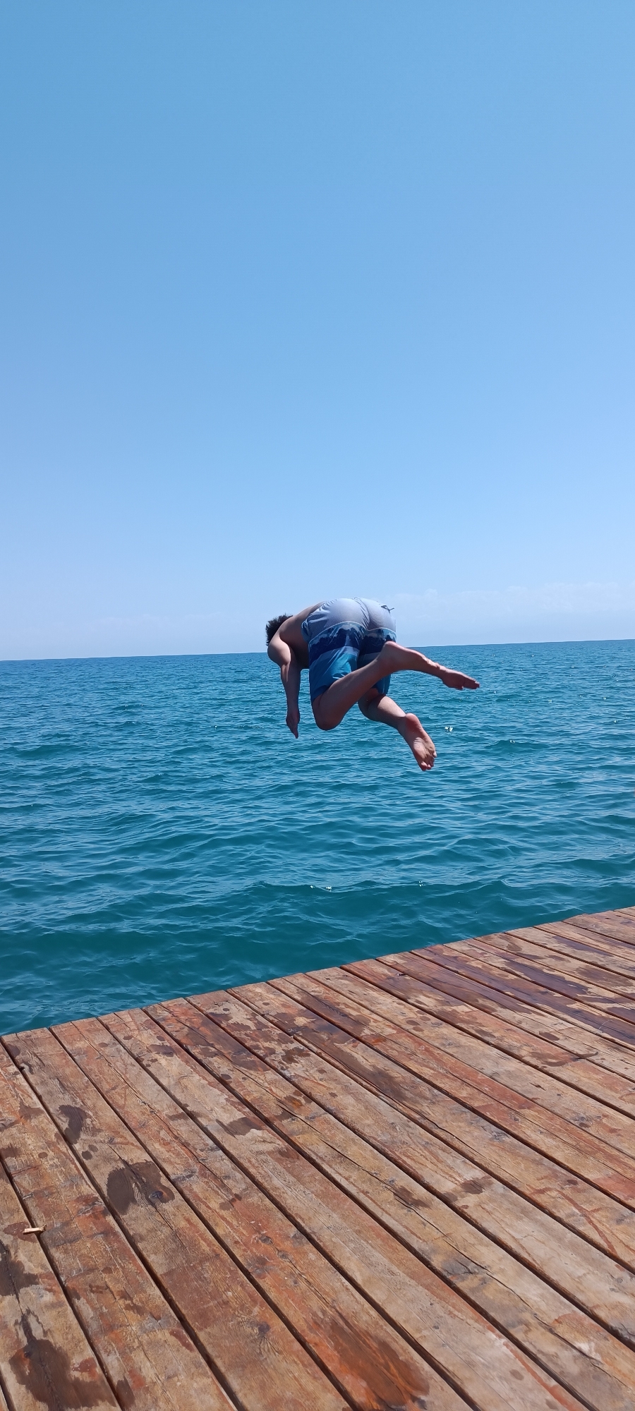 A person is captured in mid-air as they execute a dive into the blue, choppy water from a wooden dock. The person, wearing blue patterned shorts and a light blue shirt, is suspended horizontally, with their body arched and legs extended. Their face is obscured as they are facing downwards. The wooden dock, made of weathered planks, dominates the foreground of the image, showing signs of water saturation. The background consists of a clear, cloudless blue sky meeting the expansive ocean horizon. The setting appears to be an outdoor recreational area, likely a lake or sea, under bright daylight conditions. The location is Unknown, Unknown.