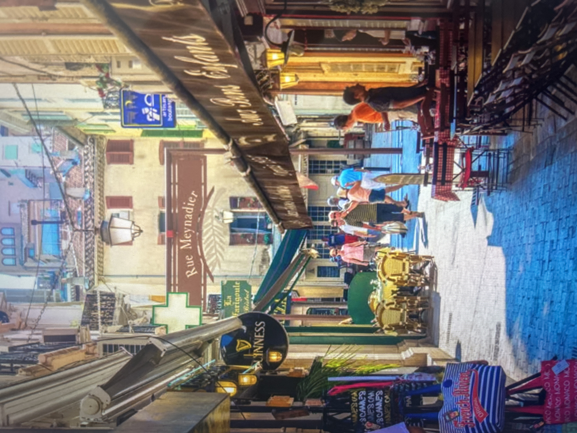 The image captures a vibrant, narrow street lined with multi-story buildings, shops, and restaurants, indicating a bustling commercial or tourist area. The street, paved with a patterned or cobblestone surface, is seen looking down its length, filled with activity.
Several people are visible, walking or standing, and some appear to be enjoying outdoor dining at tables arranged along the street. These tables, some featuring red and white checkered tablecloths, along with chairs, create inviting seating areas. The buildings display traditional architecture, with various window styles and awnings extending over the street for shade. Street lamps and other light fixtures contribute to the ambiance.
Numerous signs help identify the establishments. A prominent circular black sign with white text clearly displays "GUINNESS". A blue rectangular sign indicates an "artisan boulang" (artisan bakery). A brown banner sign hanging across the street boldly reads "RUE MEYNADIER", identifying the specific street. Another long, wooden sign above an awning says "Chez les Enfants". A green sign partially shows "La Farigoule Bistrot".
Shopping bags, including one with blue and white stripes, feature text such as "CANNES", "MONACO", and "French Riviera", along with repeated mentions like "SCANNES" and "MONICO MONA". These textual cues strongly suggest the location is in the French Riviera, likely Cannes. The scene unfolds during daylight hours, under clear skies, with a pleasant mix of sun and shade.