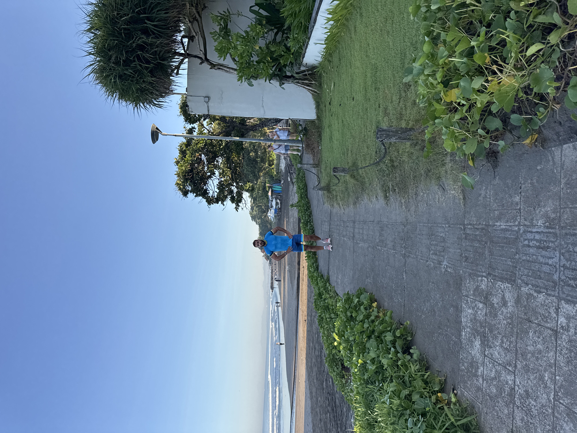 A man stands on a concrete path near a beach in Kuta, Indonesia. He is wearing a blue shirt and blue shorts with pink shoes. The path is lined with green vegetation. To the left of the path is a beach and the ocean. Other people are visible in the background. The sky is clear blue.