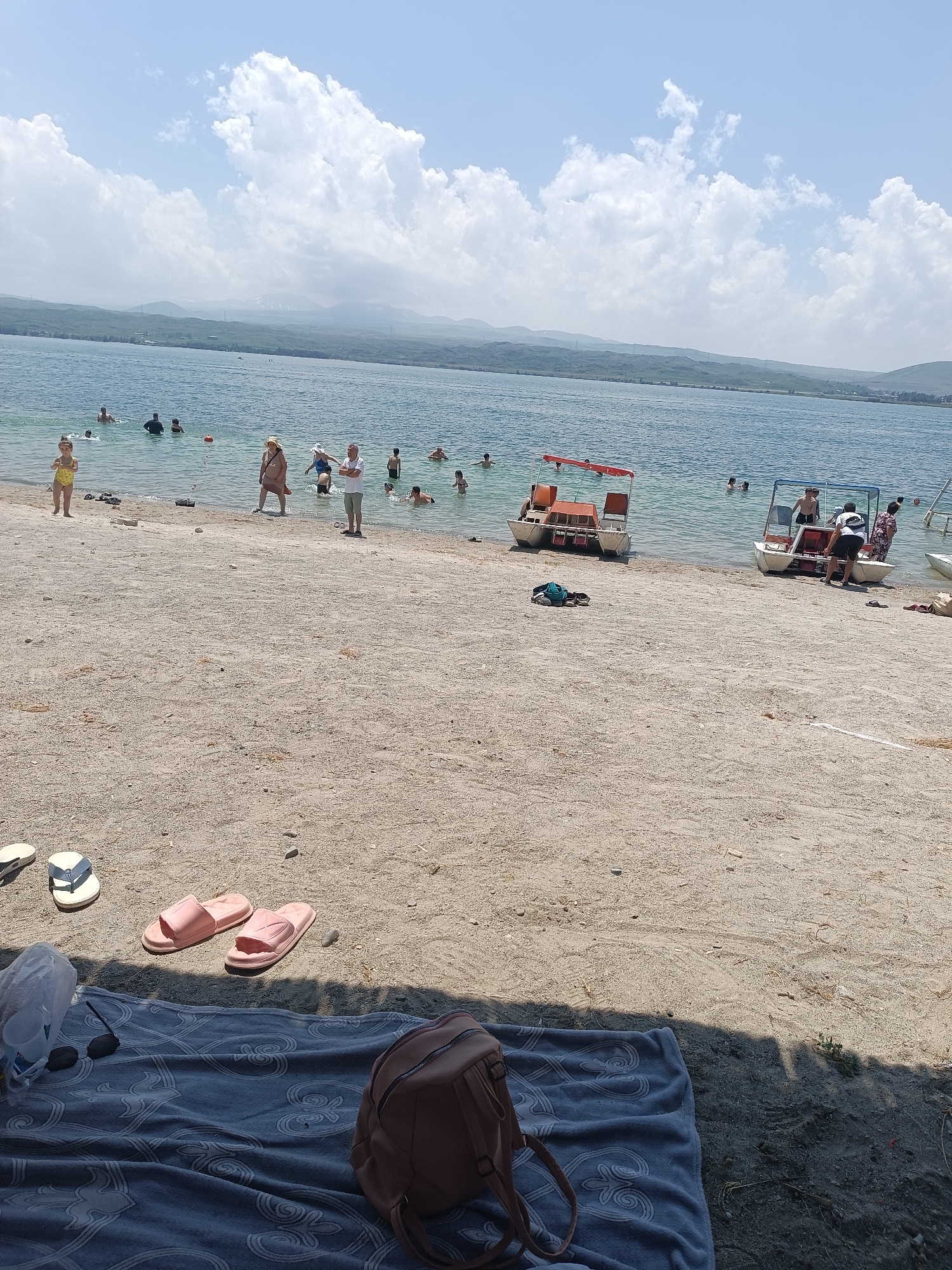 A sunny day at the beach in Sevan, Armenia. The scene captures a section of the shore of Lake Sevan with several people enjoying the water and the sandy beach. In the foreground, a grey patterned towel is spread on the sand, with a pink backpack, a plastic bag, sunglasses, and a pair of pink flip-flops resting on it. Two other pairs of flip-flops are visible on the sand further up the beach.
The water is calm and clear, with several individuals swimming and wading. A small red and white pedal boat is docked on the right side of the image, with people boarding or disembarking. Another similar pedal boat is positioned further to the left. The background shows a line of green hills and mountains under a partly cloudy sky, typical of the Sevan landscape. The overall atmosphere is one of leisure and recreation on a warm summer day.