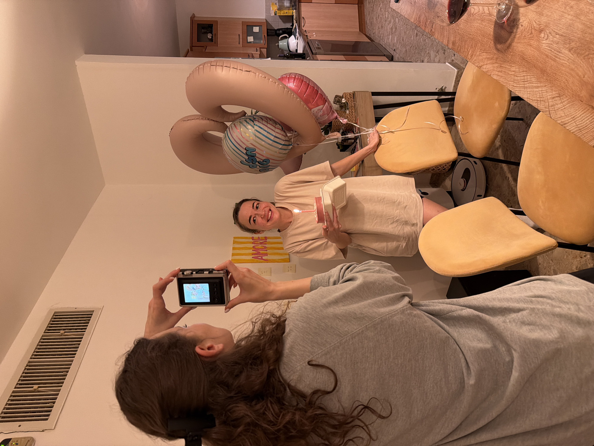 A photograph taken indoors shows two women celebrating a birthday. One woman holds a small birthday cake with a lit candle and balloons that spell out the number "36". She is smiling and looking at the camera. The other woman is holding a small digital camera and taking a photo of her. They are in a room with a dining table and chairs, and a kitchen is visible in the background. A yellow and orange painting is visible on the wall. The location is potentially a private residence, but there is no clear evidence that is in Rishon LeZion, Israel.