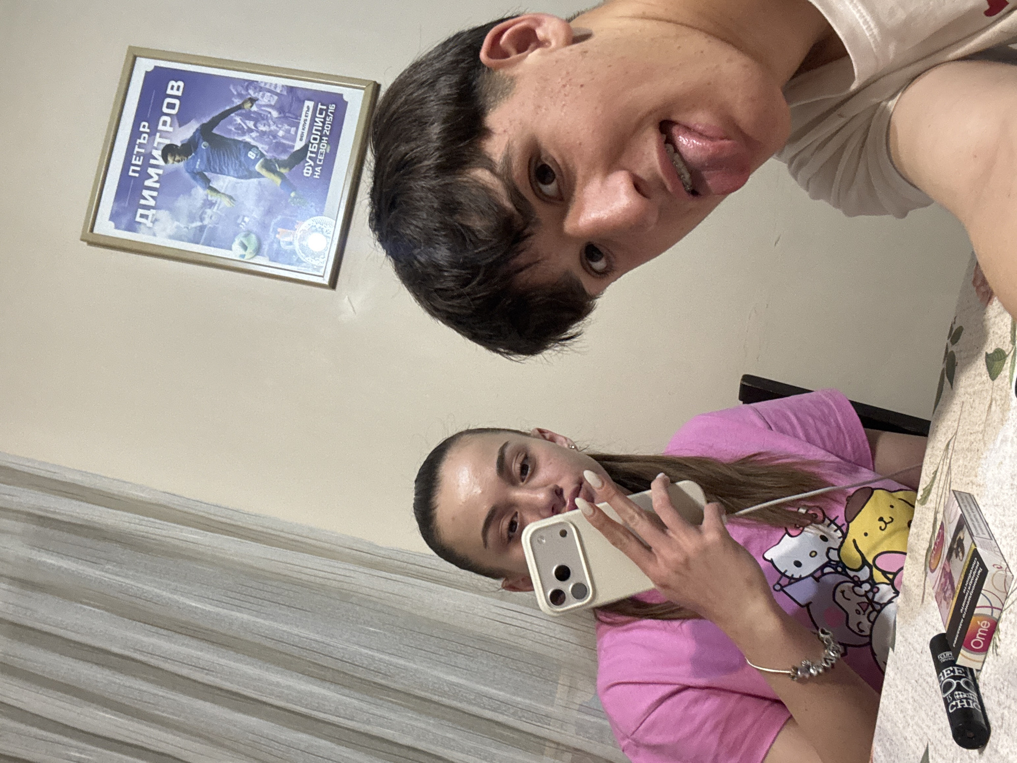 Two individuals are visible in the image, a male and a female, both appearing relatively young. The male is positioned at the top of the frame, showing his face with his tongue sticking out. The female is visible in the lower portion of the frame, holding a phone and appearing to take a selfie. She is wearing a pink shirt with a cartoon print. A pack of "Ome" cigarettes and a lighter are placed on the table. A framed poster featuring a soccer player named "Petar Dimitrov" is hung on the wall behind them. The environment shows a room with curtains and a table. Location information: The poster indicates that Petar Dimitrov is a soccer player from Bulgaria, and the cigarette brand and text on the poster use the Bulgarian language. The city name "Bozhurishte" is based on image content. Therefore, the picture seems to have been taken in Bozhurishte, Bulgaria.