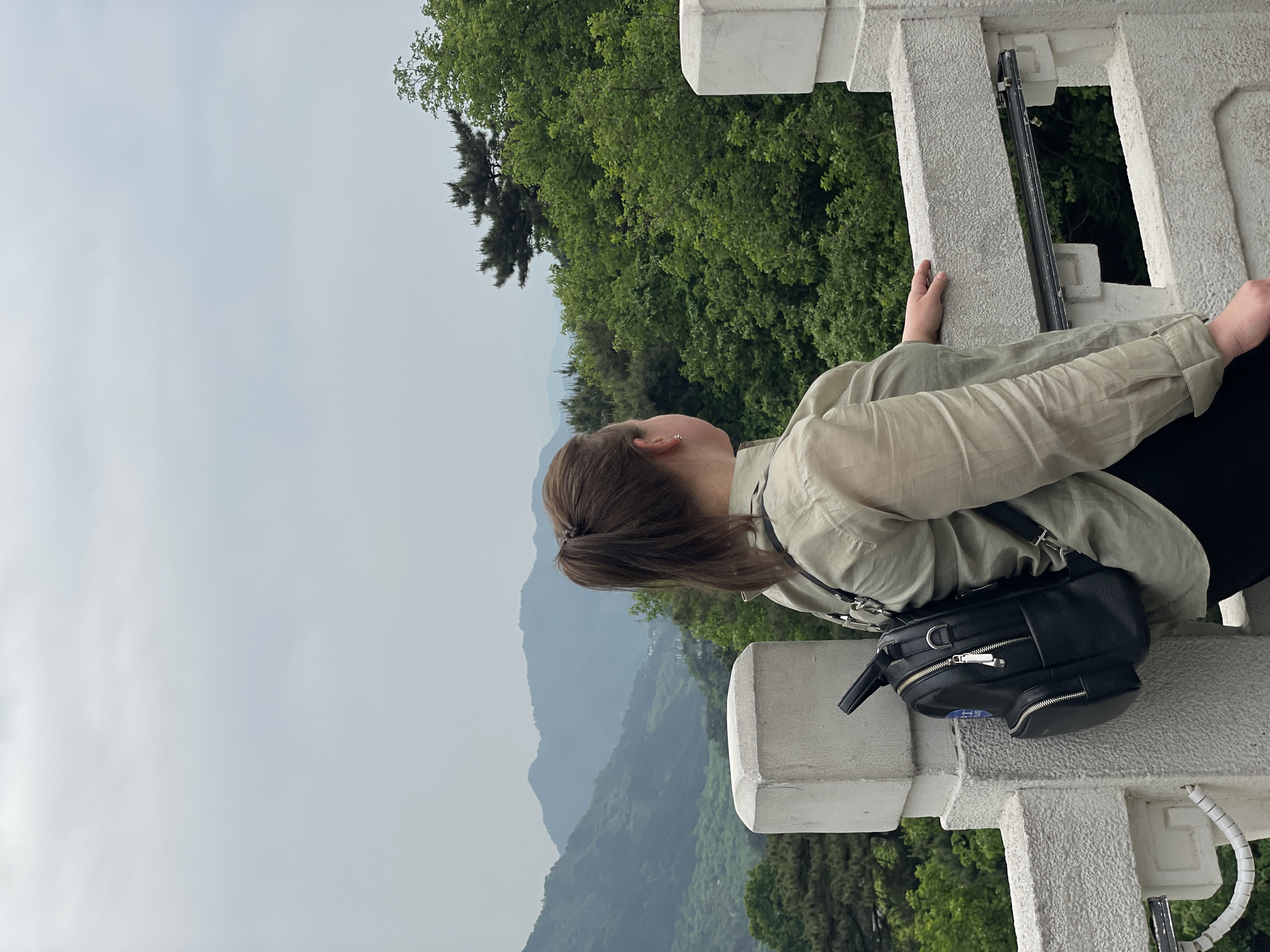 A woman is leaning over a white stone balustrade looking out over a mountainous landscape in Huairou District, China. She wears a light-colored button-down shirt, dark pants, and a black leather bag. Her brown hair is pulled back. The sky is overcast. Green trees and misty mountains fill the background.