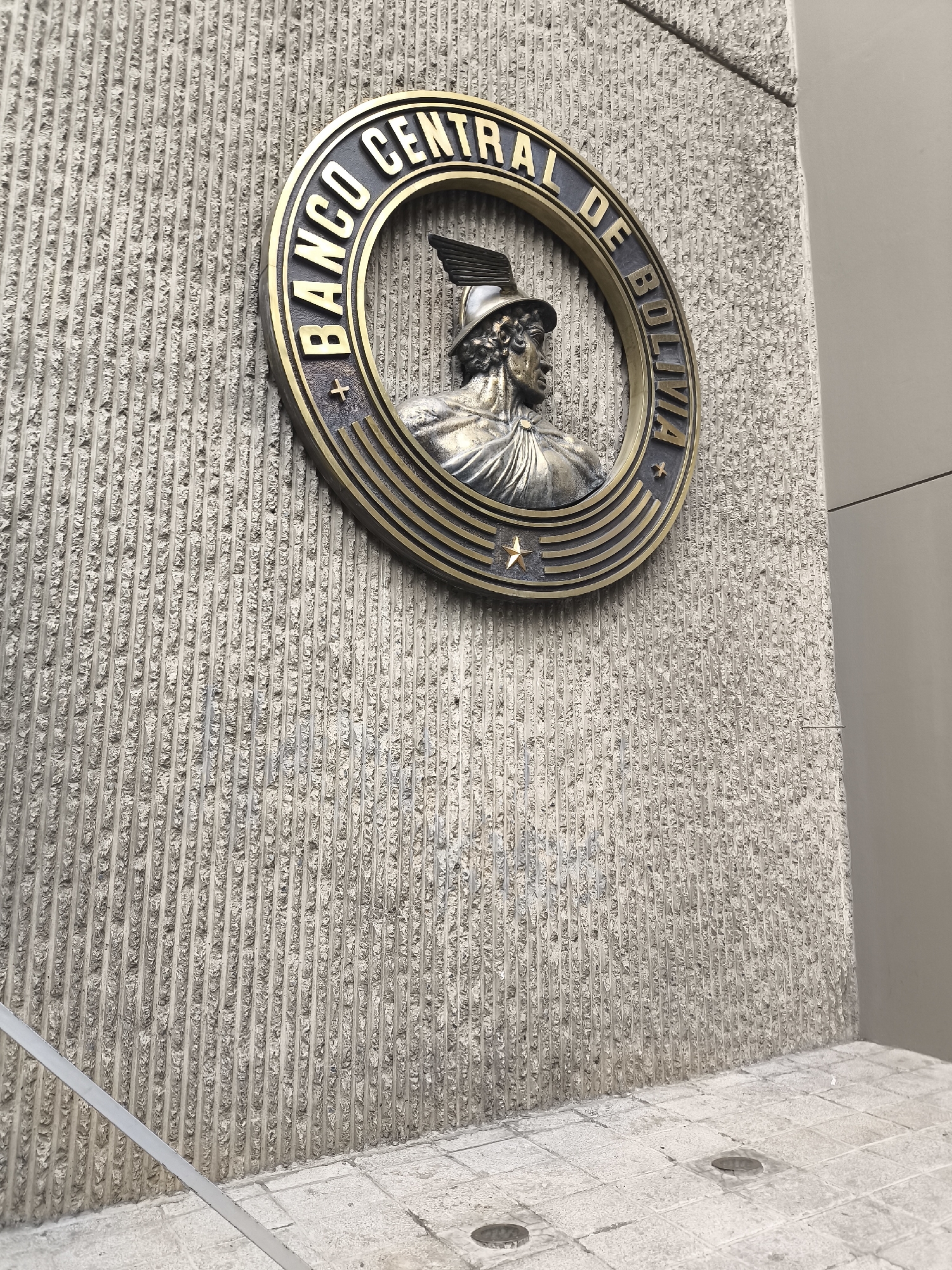 A circular emblem is mounted on a textured concrete wall. The emblem is bronze and features the words "BANCO CENTRAL DE BOLIVIA" in gold lettering around its perimeter. In the center of the emblem is a relief sculpture of a bust of a person wearing a helmet with wings and a draped garment. The emblem is positioned at an upward angle. The concrete wall has a rough, ridged texture, and a light-colored railing runs diagonally across the lower left portion of the image. The ground in the foreground consists of light-colored square tiles. The scene appears to be an exterior view of a building.