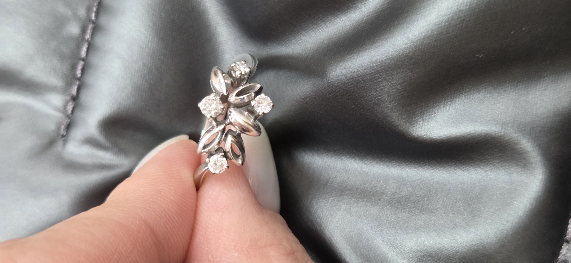A close-up, eye-level shot features a person's hand holding a ring against a dark, lustrous fabric background. The hand, with light-colored nail polish on the visible fingers (thumb and index finger), gently grasps the band of the ring.
The ring is silver-toned and intricately designed with a floral or leafy motif. It showcases several stylized leaves and small, round elements, some solid and some open-cut. Five small, round, clear gemstones are set within this design, sparkling brightly and likely being diamonds or cubic zirconia. The metal of the ring appears polished and reflective.
The background is a dark grey or charcoal-colored fabric, possibly satin or silk, characterized by its smooth, shiny surface and numerous soft folds and wrinkles. A distinct seam with visible stitching is present on the fabric, suggesting it is a draped textile or part of a garment. The lighting is soft, highlighting the textures and shine of the jewelry and the fabric. No text is visible in the image.