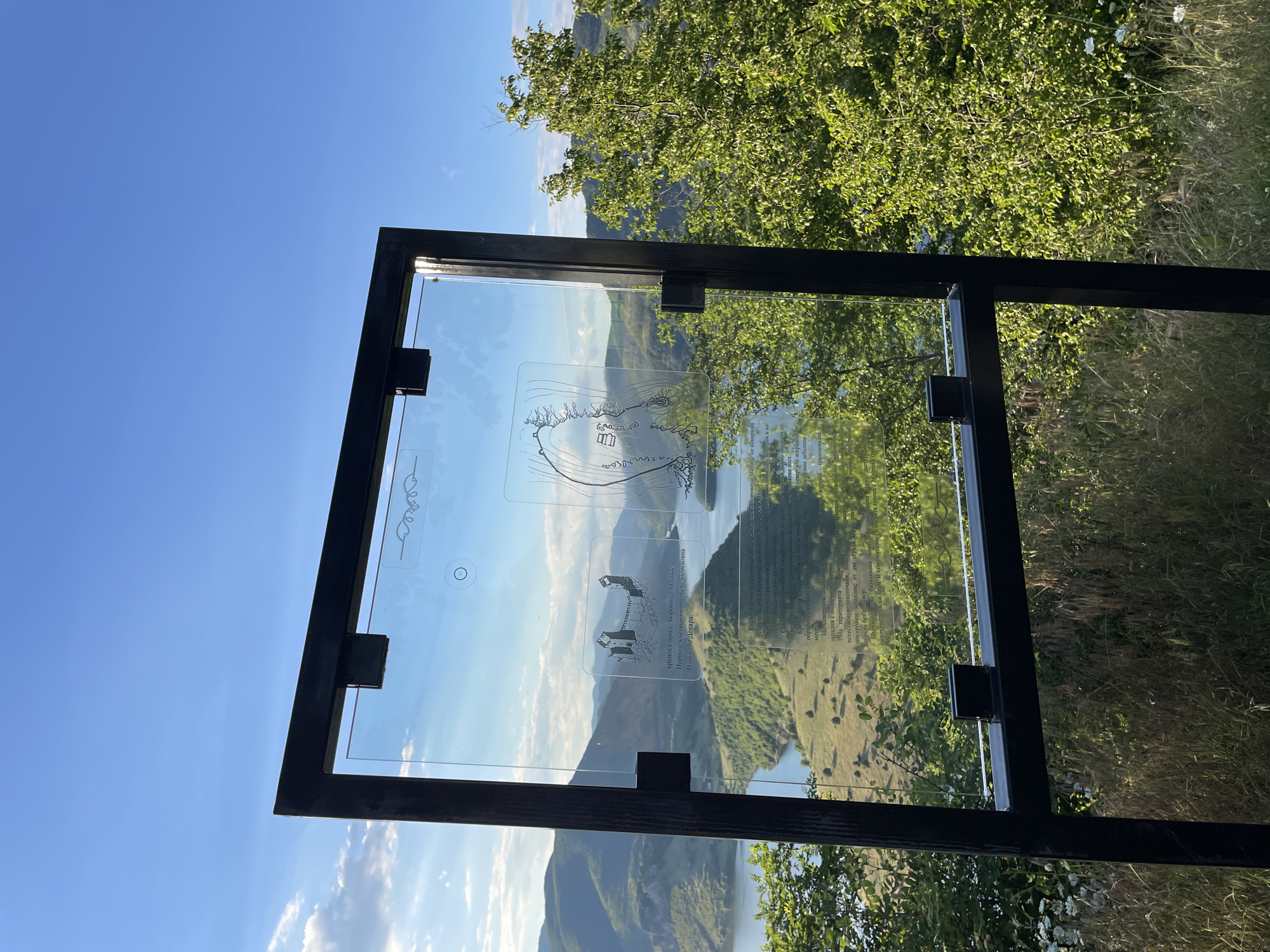A clear glass information board with a black metal frame stands on a grassy hill overlooking a large body of water surrounded by green, rolling hills under a bright blue sky with scattered white clouds. The board displays text and illustrations in Bulgarian, with a depiction of a castle or fortress on the left and a map or diagram on the right. The sun is setting, casting a warm light on the landscape. The location is identified as Ribartsi, Bulgaria.