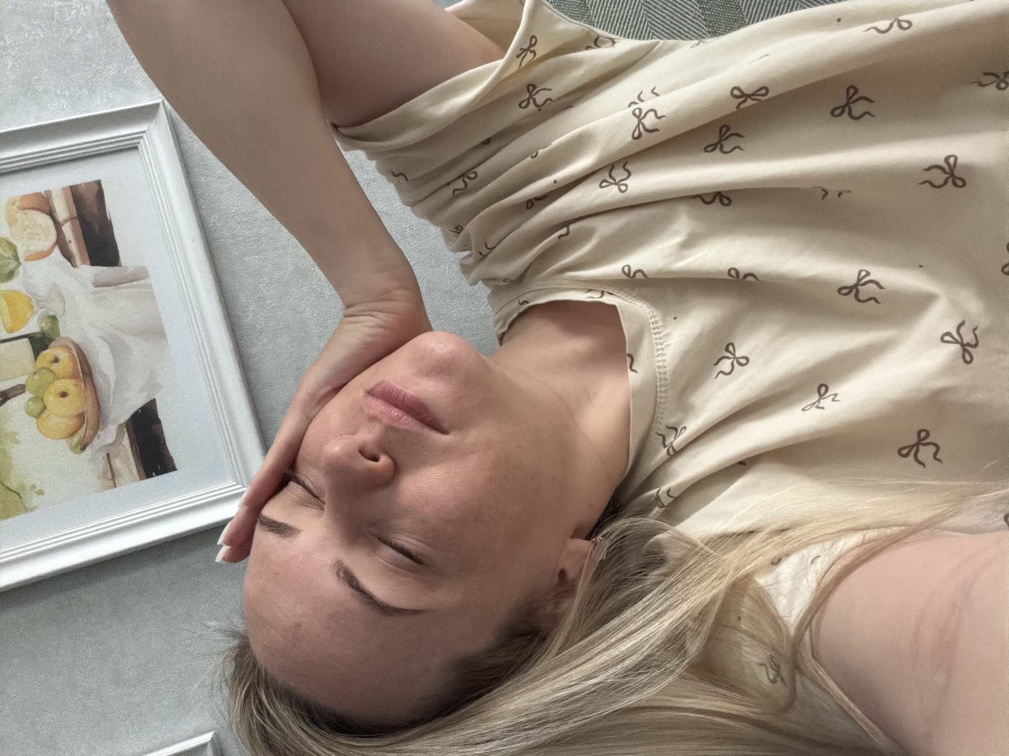 A woman with blonde hair is lying down with her eyes closed and her hand resting on her forehead. She is wearing a light-colored patterned shirt. Beside her is a framed picture on a light gray wall. The location is not discernible as Votkinsk, Russia, from the provided image.