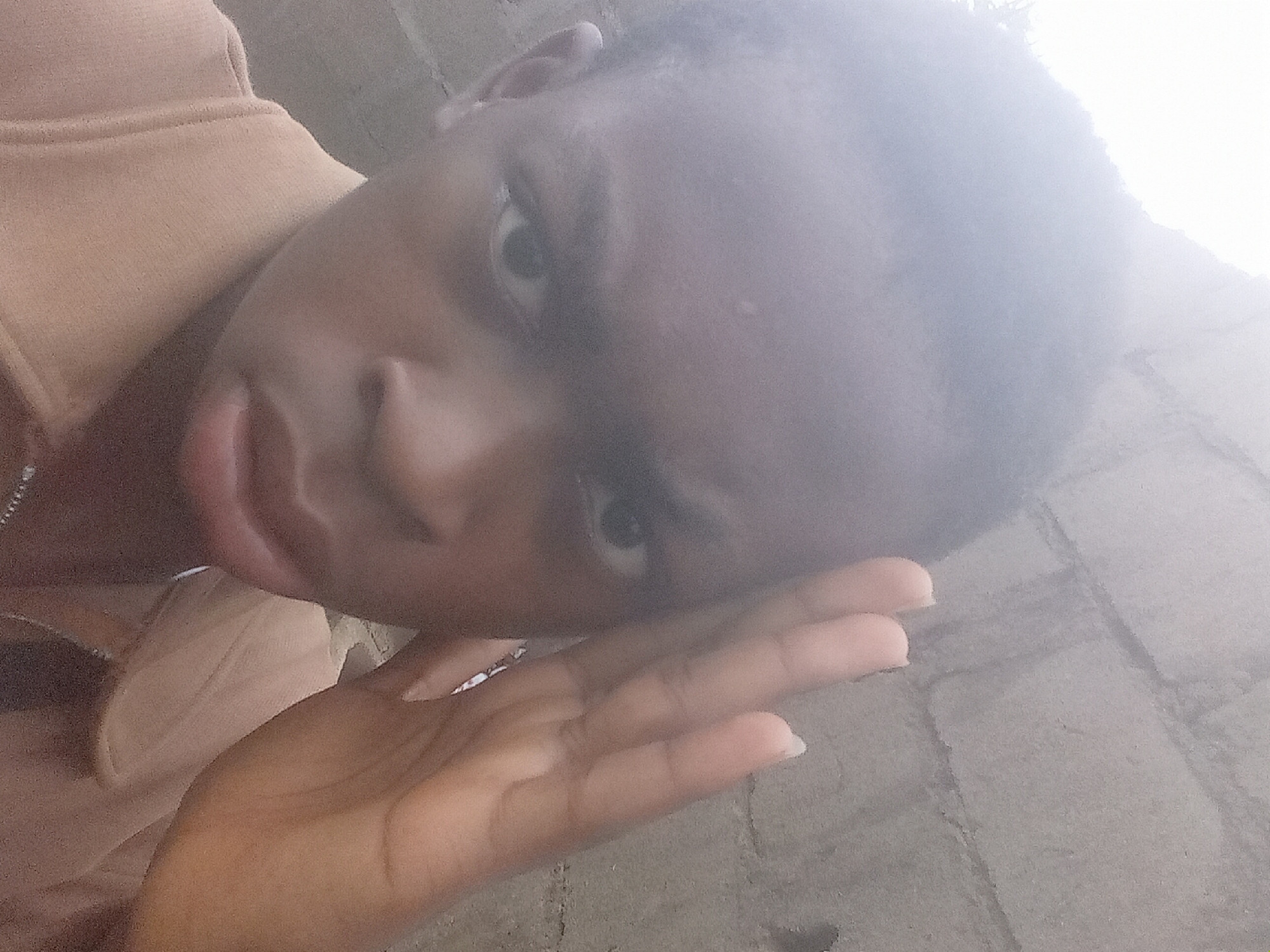 A medium, close-up shot depicts an individual with dark skin and short, dark hair. They are wearing a light-brown top. The individual's face is tilted towards the camera with their left hand resting on their face.
The background appears to be a concrete or brick surface. The location is reported to be Achara Ndiagu Omegio, Nigeria, however, no specific elements in the image confirm this location.
