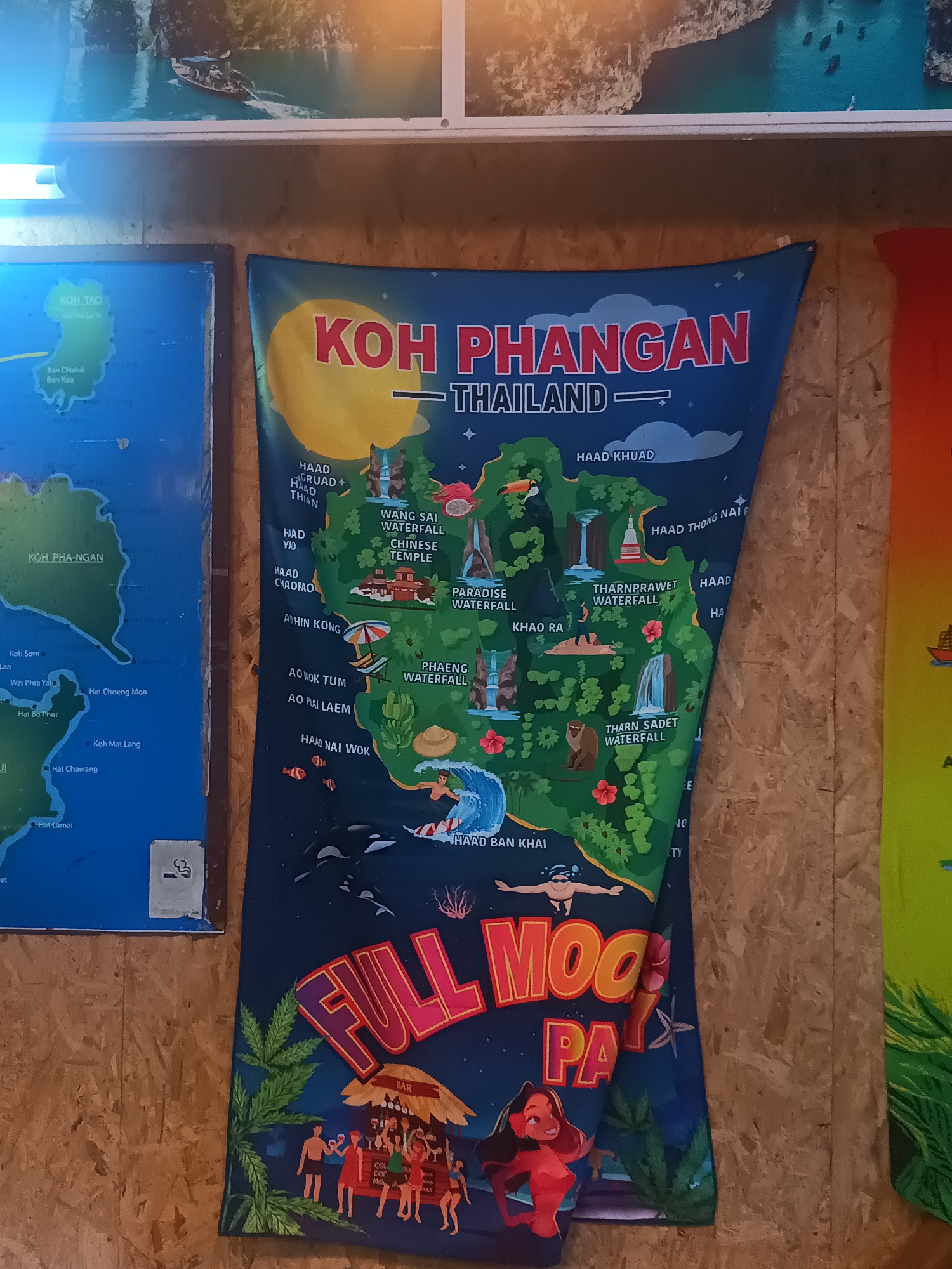 The image shows a wall with multiple posters and images. A blue map on the left features a green island labeled "KOH TAO" and another green area labeled "KOH PHA-NGAN." Next to it is a larger banner dominated by a blue background with a yellow sun-like shape. The words "KOH PHANGAN" are in large red letters, with "THAILAND" written beneath in smaller font. The banner contains illustrations of landmarks, including "WANG SAI WATERFALL," "CHINESE TEMPLE," and "PARADISE WATERFALL," among others. The bottom part of the banner features "FULL MOON PARTY" in large, vibrant letters with cartoon figures of people and marijuana leaves. The setting is a wall in Surat Thani City Municipality, Thailand.