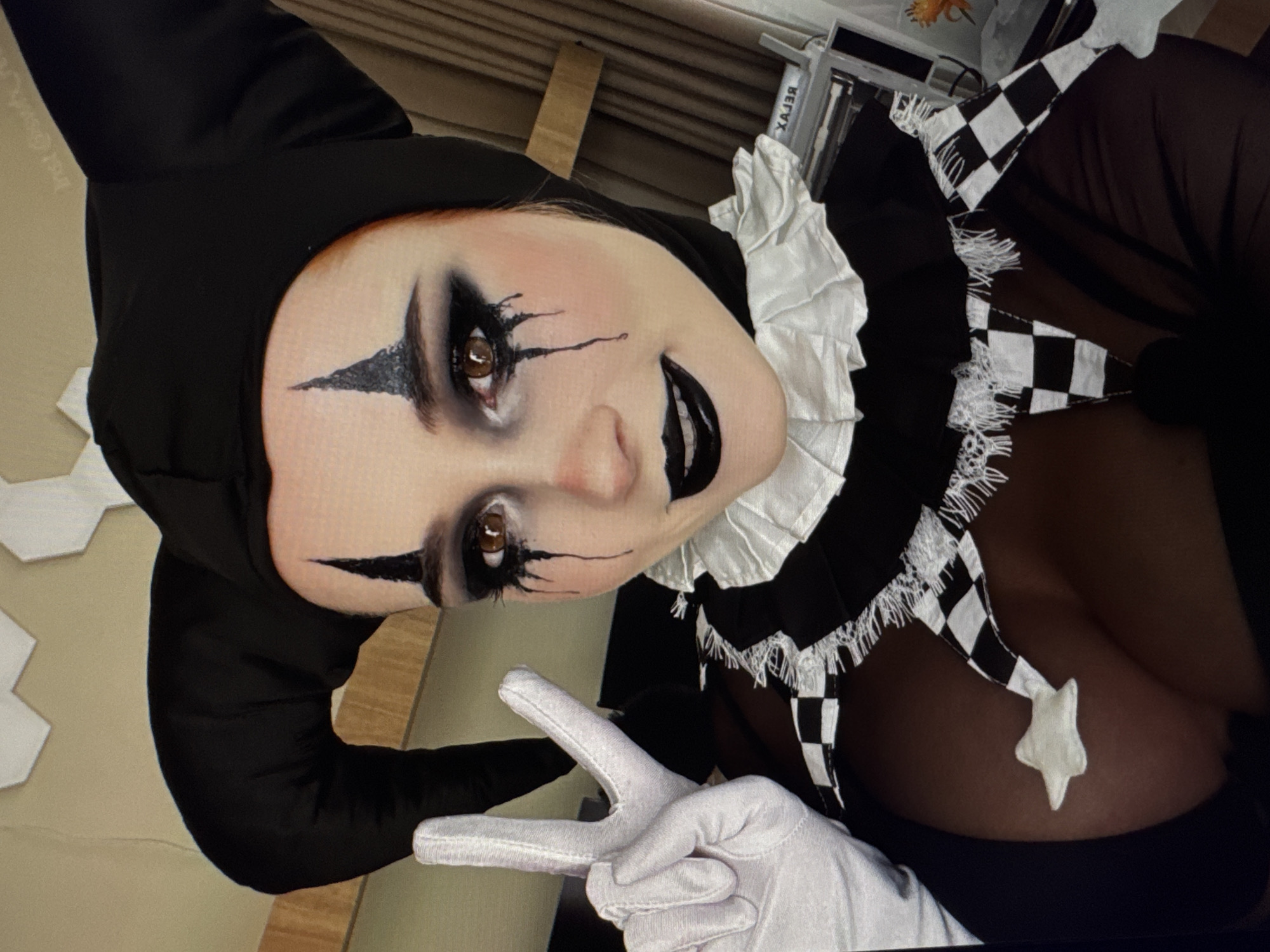 This is a close-up, angled, indoor photograph of a person dressed in a jester-like costume. The subject is wearing a black jester hat with a pointed tip, and their face is heavily made up with dramatic dark eyeshadow and eyeliner, creating a theatrical, almost harlequin-like appearance. There are also sharp, pointed black markings on their cheeks and a black, glossy lipstick. The costume features a ruffled white collar and a checkered black and white fabric detail around the neck, adorned with white fringe and a white star-shaped embellishment. The subject is wearing white gloves and is making a peace sign with their right hand. The background is out of focus, but appears to be an indoor setting with a wall and some shelving or furniture. The lighting is artificial. The overall mood of the image is theatrical and possibly playful or unsettling due to the makeup and costume. There is no visible text.