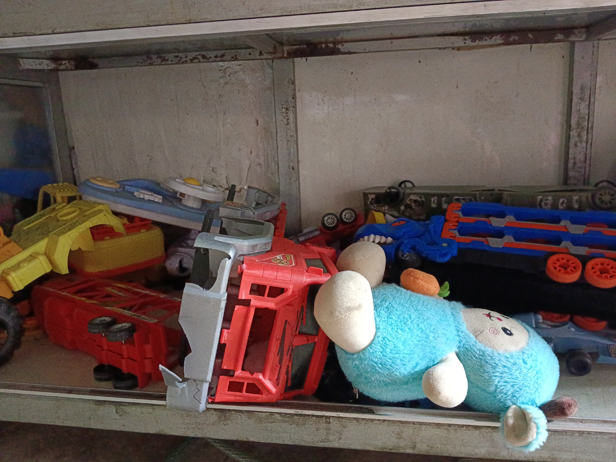 The image depicts a shelf cluttered with various children's toys. On the left, a yellow toy truck is partially visible. Next to it, a red toy truck is overturned, with its wheels facing upwards. A blue and white toy boat with yellow accents is also on the shelf. In the center, a red toy vehicle chassis is visible. To the right of the red chassis, a light blue stuffed animal with white stuffed limbs is lying down, with an orange carrot-shaped toy near its head. Behind the stuffed animal, a blue and orange toy train or vehicle is present. The shelf appears to be made of metal and is set against a textured, light-colored wall. The overall environment suggests a storage area or a play area that is not currently in use. The location is specified as Aek Kanopan, Indonesia.