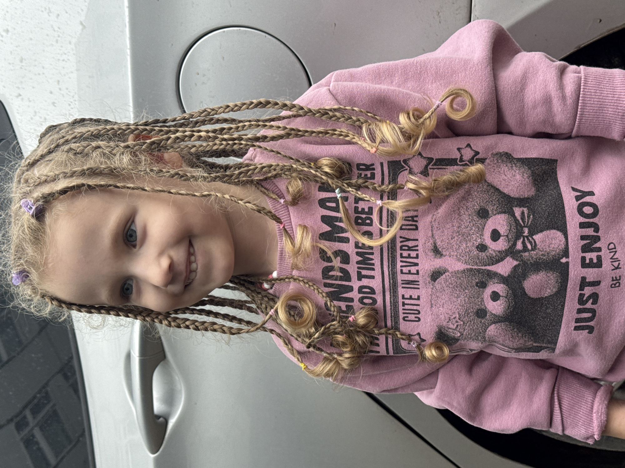 A young girl with blonde hair styled in numerous thin braids adorned with small colorful hair ties, some of which are coiled, stands in front of a silver car. She is wearing a pink sweatshirt with a graphic print of two teddy bears and text that reads "FRIENDS MAKE GOOD TIMES BETTER" and "CUTE IN EVERY DAY JUST ENJOY BE KIND." The background consists of the car and an overcast sky, suggesting it might be a slightly damp day as evidenced by water droplets on the car's surface. The scene appears to be in an urban or suburban setting.