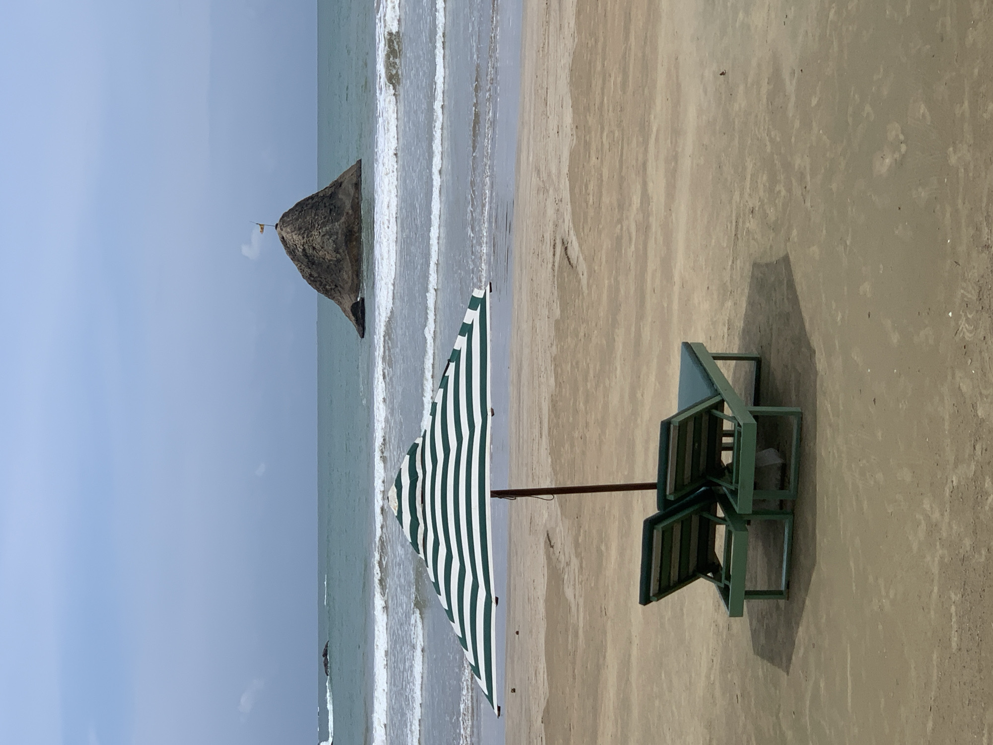 The image depicts a coastal scene in Weligama, Sri Lanka. Two unoccupied, green wooden beach chairs are positioned on a sandy beach under the shade of a green and white striped umbrella. Beyond the chairs and umbrella, the ocean stretches out with gentle waves breaking on the shore. In the distance, a large, rocky formation rises from the sea. The sky is overcast.
