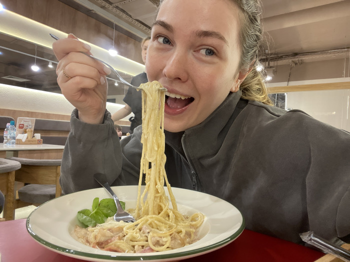 A blonde-haired woman enthusiastically eats a forkful of spaghetti carbonara. She's indoors, presumably in a restaurant, with a full plate of pasta in front of her. The spaghetti is topped with parmesan cheese, and fresh basil leaves garnish the dish. A second fork rests on the plate. The table is red, and water bottles and other restaurant elements are visible in the blurred background. The woman is wearing a zip-up sweatshirt.