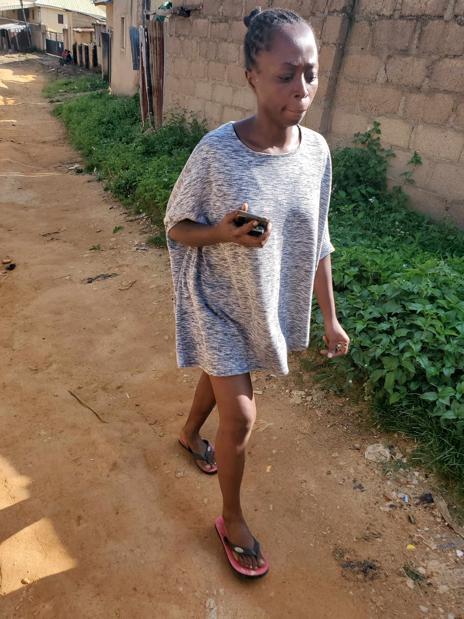 A young Black woman with her hair pulled back in a bun walks on a dirt path, looking down at her cell phone. She is wearing a grey and white patterned, oversized shirt and pink and black flip-flops. The path is lined with green vegetation on the right side and buildings on the left. In the background, there are buildings that appear to be in a state of disrepair, with corrugated metal roofs and brick walls. The scene is likely located in Jos, Nigeria, given the architectural style and vegetation.