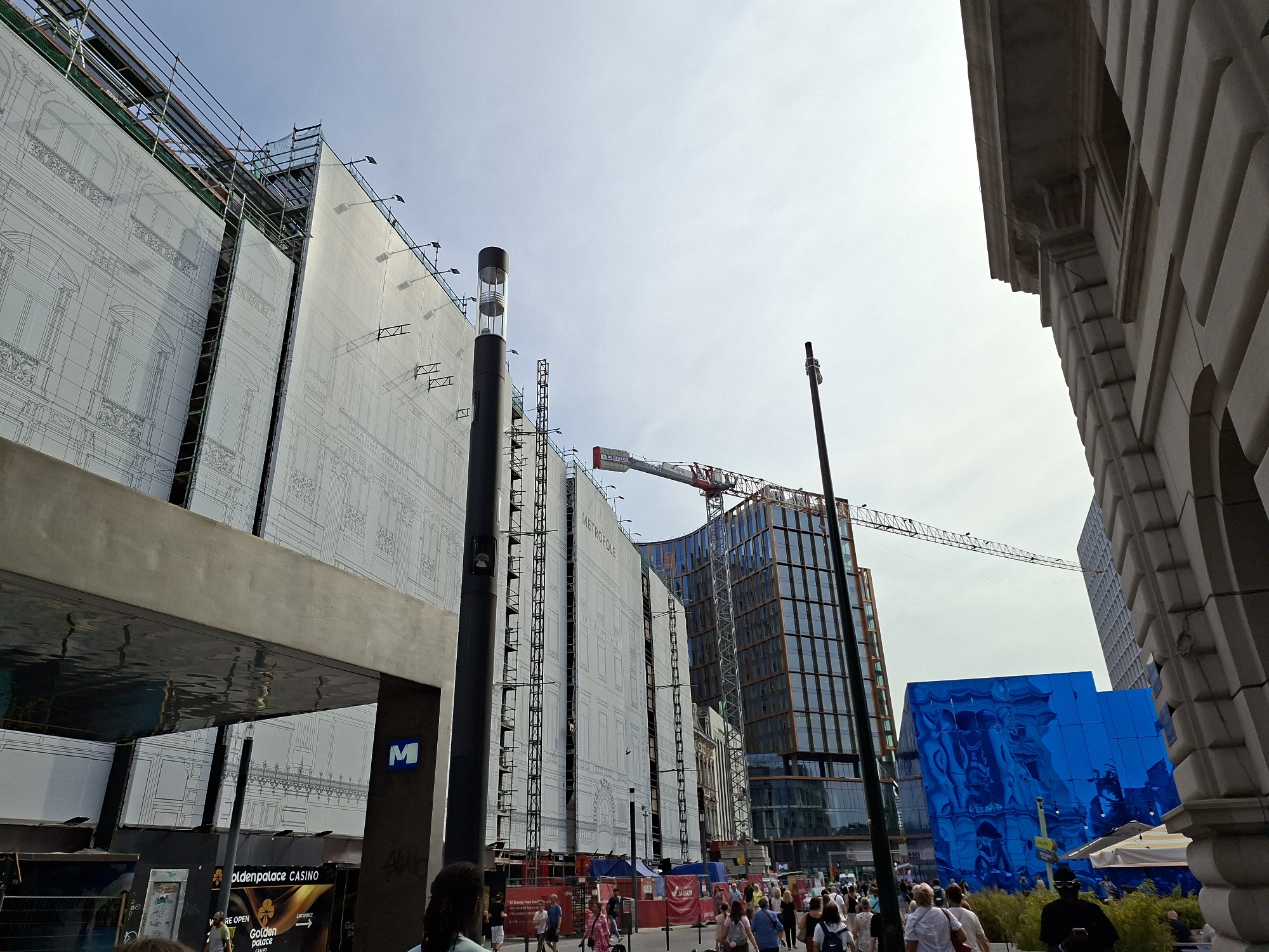This is a street-level, eye-level photograph taken on a cloudy day in Brussels, Belgium. The scene depicts a busy urban environment with ongoing construction and a mix of modern and older architecture. In the foreground, to the left, is a reflective ceiling of a modern structure, with its surface rippling as if mirroring the environment. Below this, a section of a casino signage is visible, reading "goldenpalace CASINO". A dark, cylindrical lamppost stands to the right of the reflective ceiling.
Dominating the left and center of the image are large white banners draped over a building undergoing renovation. These banners are printed with the facade of a classical building, complete with detailed architectural drawings of windows and decorative elements. Scaffolding and construction equipment are visible behind and around these banners. To the right of these banners, a modern, glass-fronted building rises, with its reflective surface showing the sky and surrounding structures. A construction crane is visible extending from behind this building and reaching across the sky.
Further to the right, a striking, angular building covered in reflective blue panels stands out, distorting the reflections of its surroundings. A dark, slender lamppost is positioned in front of this blue building. In the background, a crowd of people are walking along a sidewalk, suggesting a public thoroughfare. The sky is overcast with a bright, diffused light. The overall impression is one of urban development and activity.