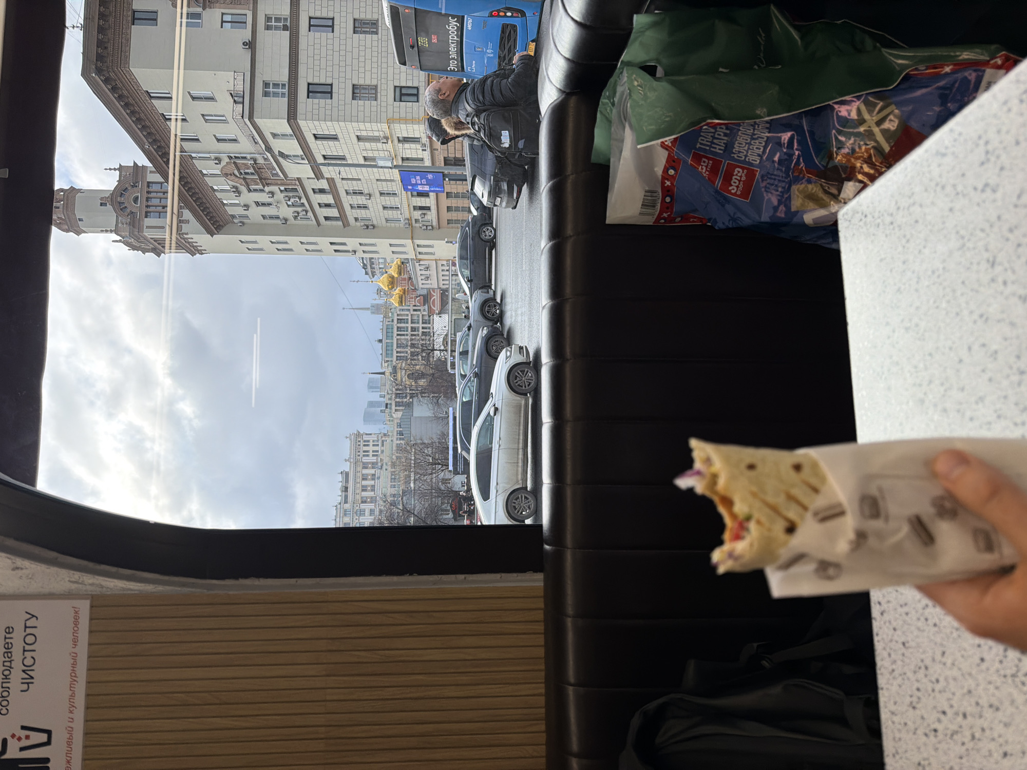Here is a detailed description of the image:
**Scene:** The image appears to have been taken from inside a bus or similar public transportation vehicle. The viewpoint faces a window, revealing an urban street scene. An arm is holding a half-eaten wrap.
**Location:** The street view shows that it is in Moscow, Russia with an Orthodox church identifiable by its multiple golden domes and the architecture of the buildings in the background.
**People:**
* One person is visible, an arm holding a half-eaten wrap.
* A second person is outside, a bus rider or pedestrian is visible next to the bus.
**Objects:**
* A partially eaten wrap
* The window and interior of a bus (seats, panels)
* Buildings: A tall building with classic architectural details, along with other buildings
* Cars parked on the street
* Shopping bags
**Environment:**
* The weather appears to be overcast.
* The location is an urban street in Moscow, Russia, characterized by multi-story buildings, cars, and an Orthodox church in the distance.