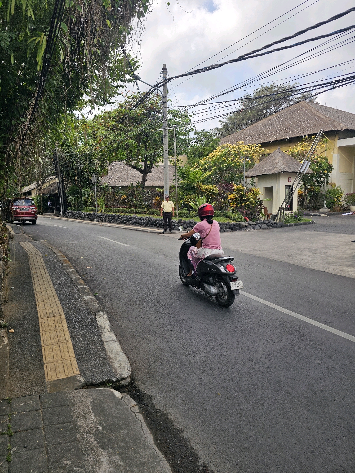 A red SUV is parked on the left side of a paved road in Denpasar, Indonesia. The road is lined with a textured yellow sidewalk and then a stone wall with lush vegetation behind it. A man in a yellow uniform stands on the sidewalk near a small building with a thatched roof. In the foreground, a woman wearing a pink shirt and patterned skirt rides a black scooter. Overhead, a dense network of electrical wires crosses the sky, partially obscuring a cloudy day. Behind the vegetation, a building with a thatched roof and yellow exterior is visible, suggesting a tropical, possibly resort-like setting. The overall scene depicts a street in a tropical city, with a mix of urban infrastructure and natural elements.