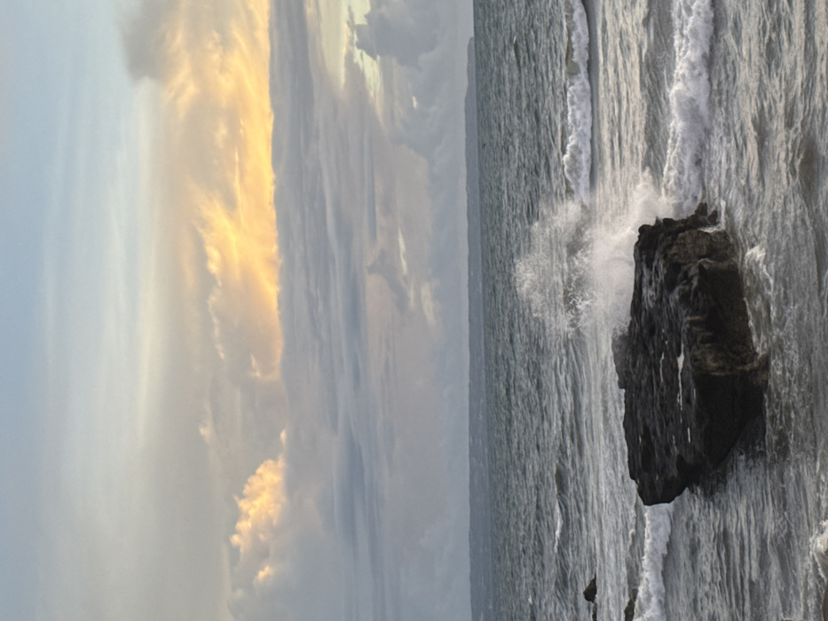 The scene depicts a turbulent coastal environment in Canggu, Indonesia, under an overcast sky with a distinct band of golden-yellow light visible within the upper cloud layers. The sky transitions from a lighter blue-grey overhead to denser, darker grey clouds towards the horizon. The ocean water is dark, choppy, and characterized by numerous waves with visible white crests. A large, dark grey, irregularly shaped rock formation is centrally positioned in the mid-foreground, partially submerged in the sea. A wave is actively crashing against the right side of this rock, generating a prominent spray of white water and foam. Smaller waves are observed both approaching the rock and further out to sea. A small, darker rock is partially visible to the left of the main formation. A low-lying landmass is faintly visible on the distant horizon, beneath the clouds. No individuals are present.