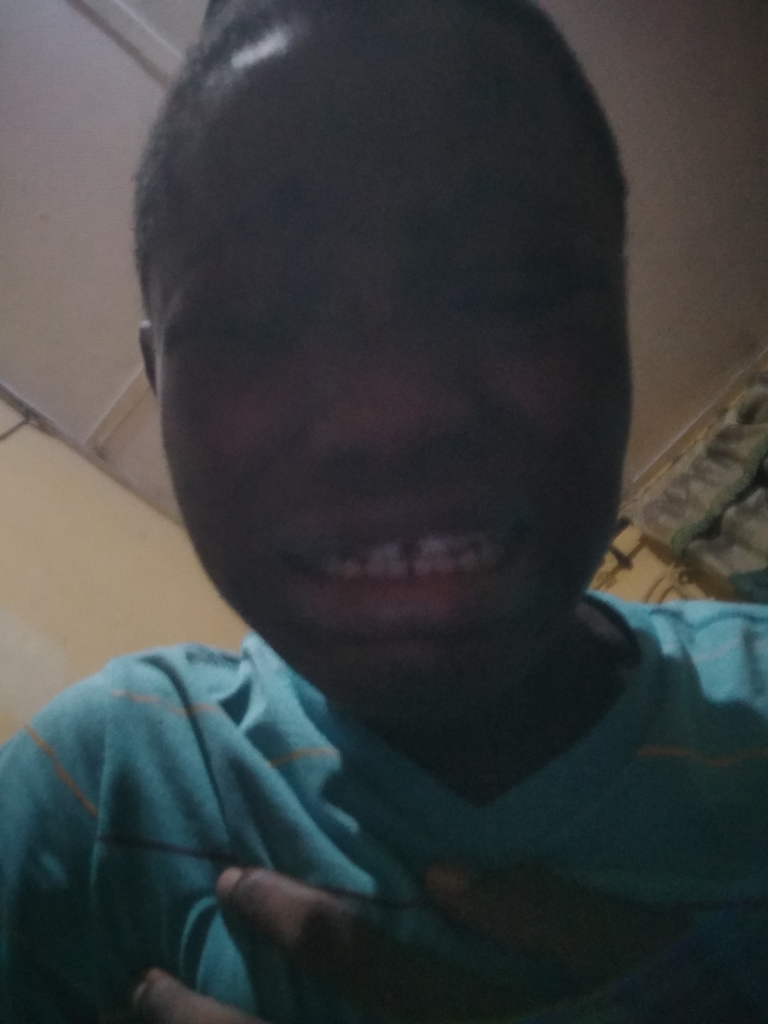 A close-up, low-angle shot shows a person with dark skin wearing a light blue t-shirt with orange horizontal stripes. The person's face is somewhat obscured by shadow. They are smiling slightly, revealing their upper teeth. One of their hands is visible at the bottom left corner of the frame. The background appears to be an indoor space with light-colored walls and a ceiling. The city and specific location are Unknown.