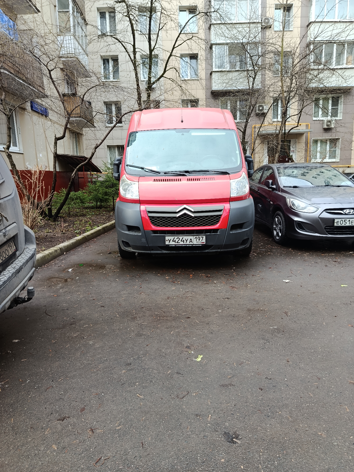 A photograph taken in Moscow, Russia shows a red Citroen van parked on an asphalt surface. The license plate, visible on the front of the van, reads "Y424YA 197 RUS." To the right of the van is a grey Hyundai Accent, with the license plate "E051EE 799 RUS." To the left of the van is a grey SUV with part of the plate "B001VV." The background includes apartment buildings with multiple windows and balconies, with bare trees in front of them. The ground is wet, and some debris is scattered on the asphalt.