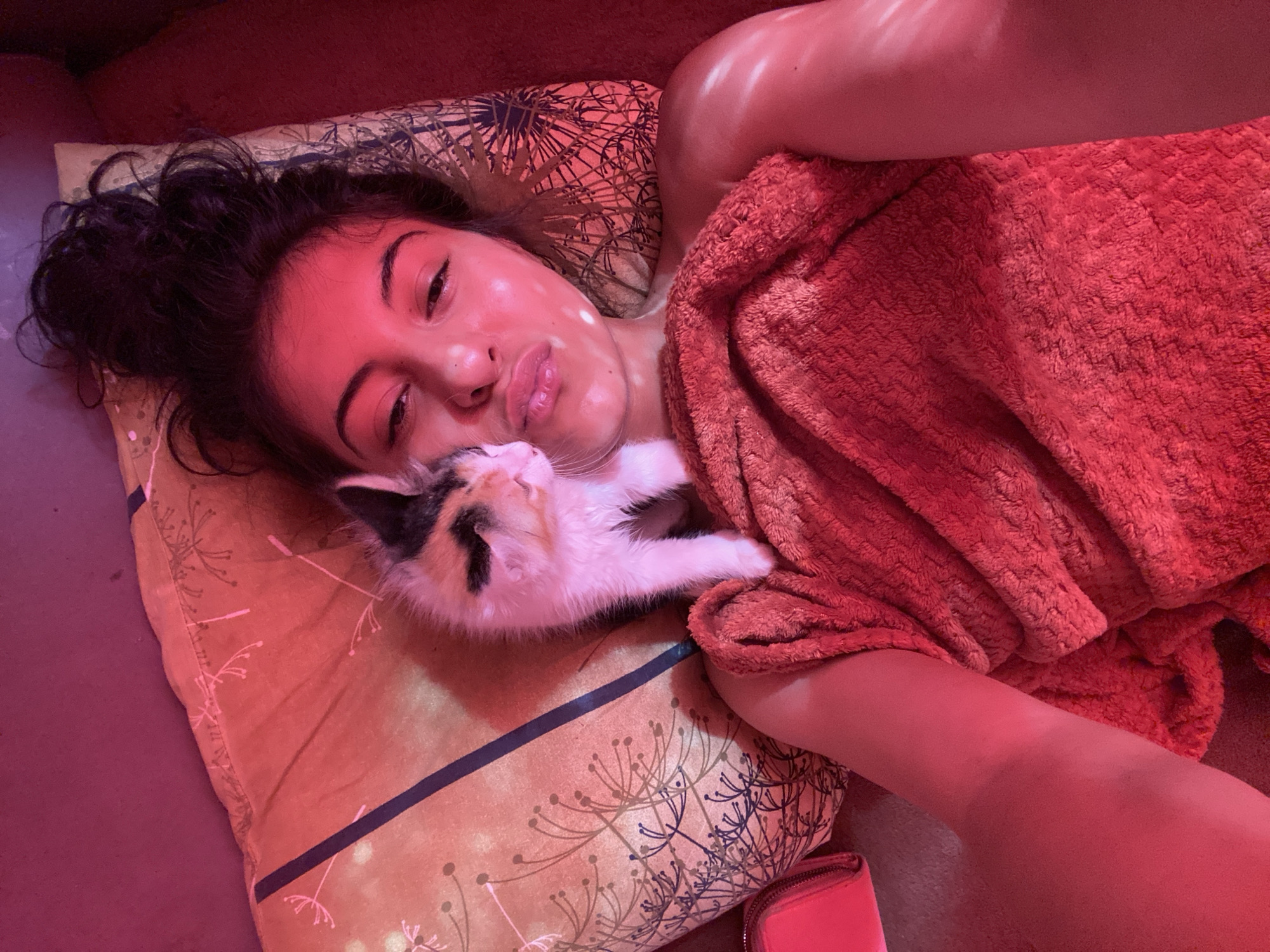 In Unknown, Unknown, a top-down, slightly angled shot captures a person, appearing female, lying supine on a patterned pillow under a dominant red ambient light. The individual's dark hair is spread on the pillow, which features a design of stylized dandelion-like plants in dark and light hues on an orange-tan base, crossed by a dark stripe. A small calico kitten, with white, orange, and black fur patches, rests on the person's upper chest/shoulder, its head turned towards and almost touching the person's nose. One of the kitten's front paws is visible on the red, fluffy blanket or towel that partially covers the person's upper body. The person's face shows full lips slightly parted, and visible highlights or reflections, possibly from a sheet mask or the lighting, are present on the forehead and left cheek. To the upper left of the frame, a bright red wallet or small pouch is visible. The immediate surroundings consist of a solid, dark reddish-brown surface.