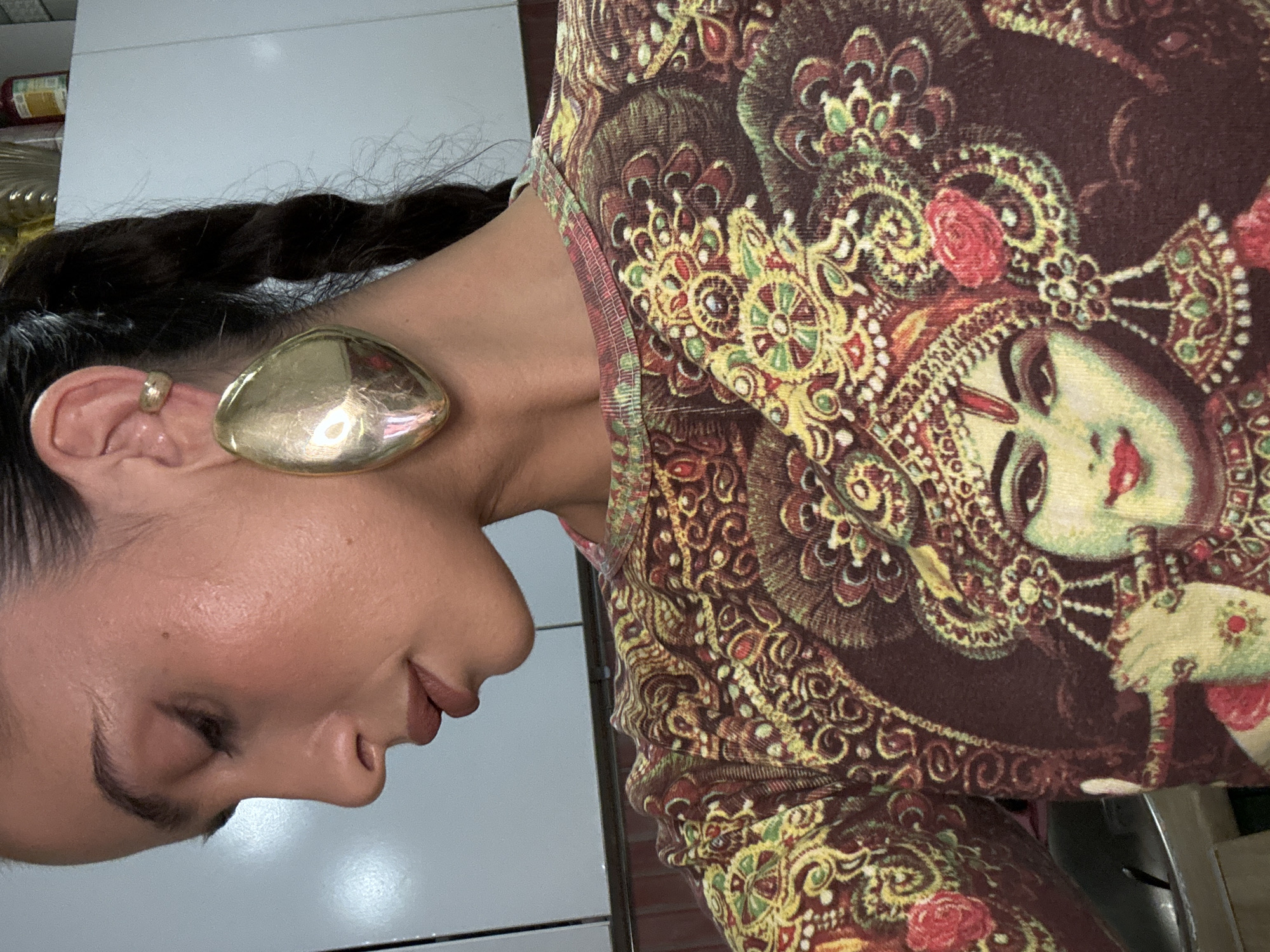 A woman is shown in a close-up shot from the left side. Her hair is styled in a braid. She has a large, shiny gold earring and a small gold ring on her ear. She is wearing a printed top depicting an ornate design with a woman's face. The background consists of a white surface, potentially a countertop. There is no indication of the location being in Krasnoyarsk, Russia.