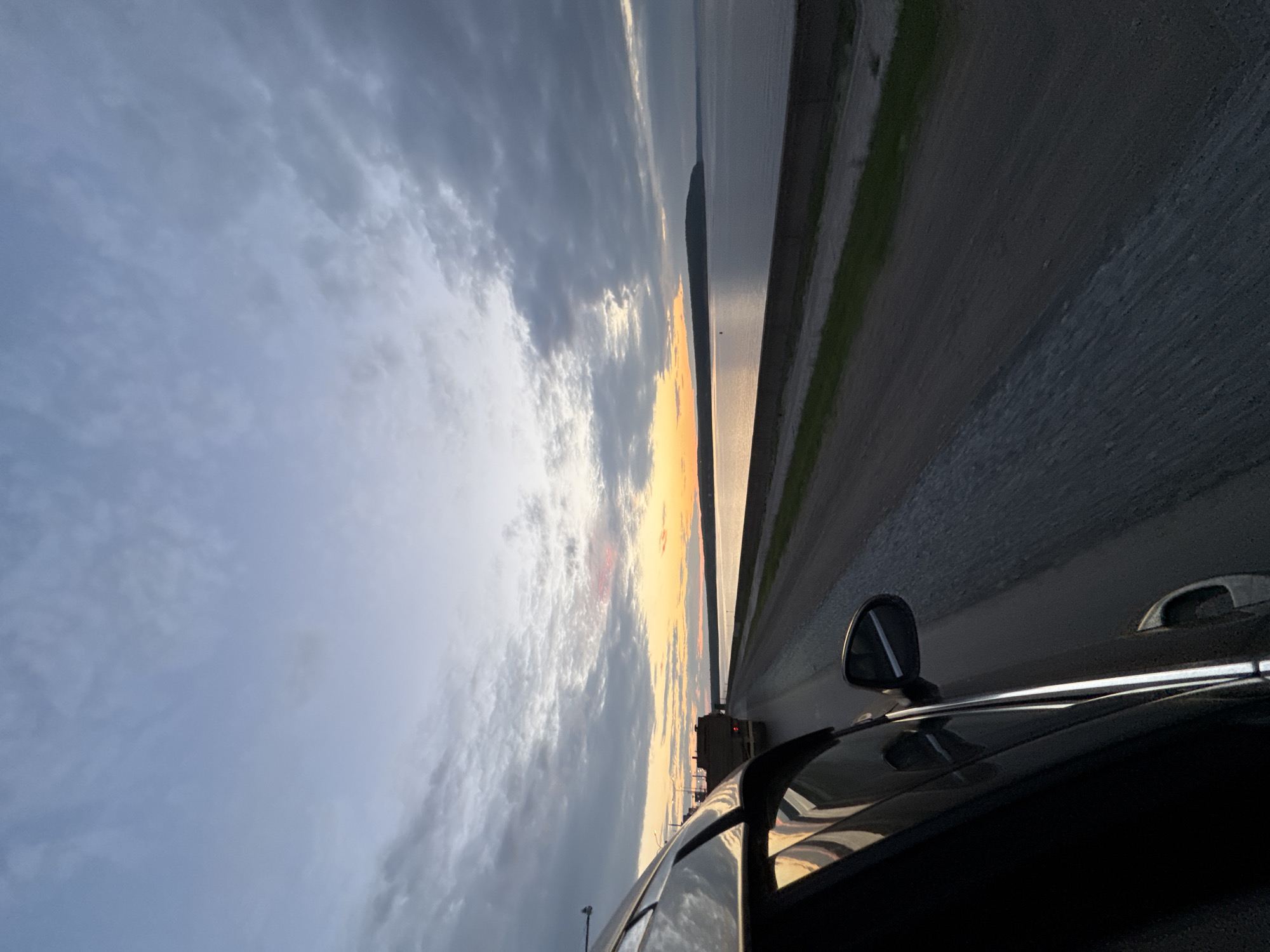 The image shows a car traveling along a coastal road at dusk. The car is dark and glossy, with its side visible from the perspective of the passenger seat. The road is paved and runs parallel to a large body of water, likely a lake or sea, with a distant, low-lying landmass on the horizon. The sky is dramatic, with a mix of dark, heavy clouds and lighter, brighter areas where the sun is setting, casting an orange glow. A truck carrying lumber is visible ahead on the road, suggesting this is a transportation route. The scene appears to be located near Noviy, Russia, given the context provided. The overall atmosphere is one of twilight travel, with the natural beauty of the landscape dominating the scene. There are no people clearly visible in the image.