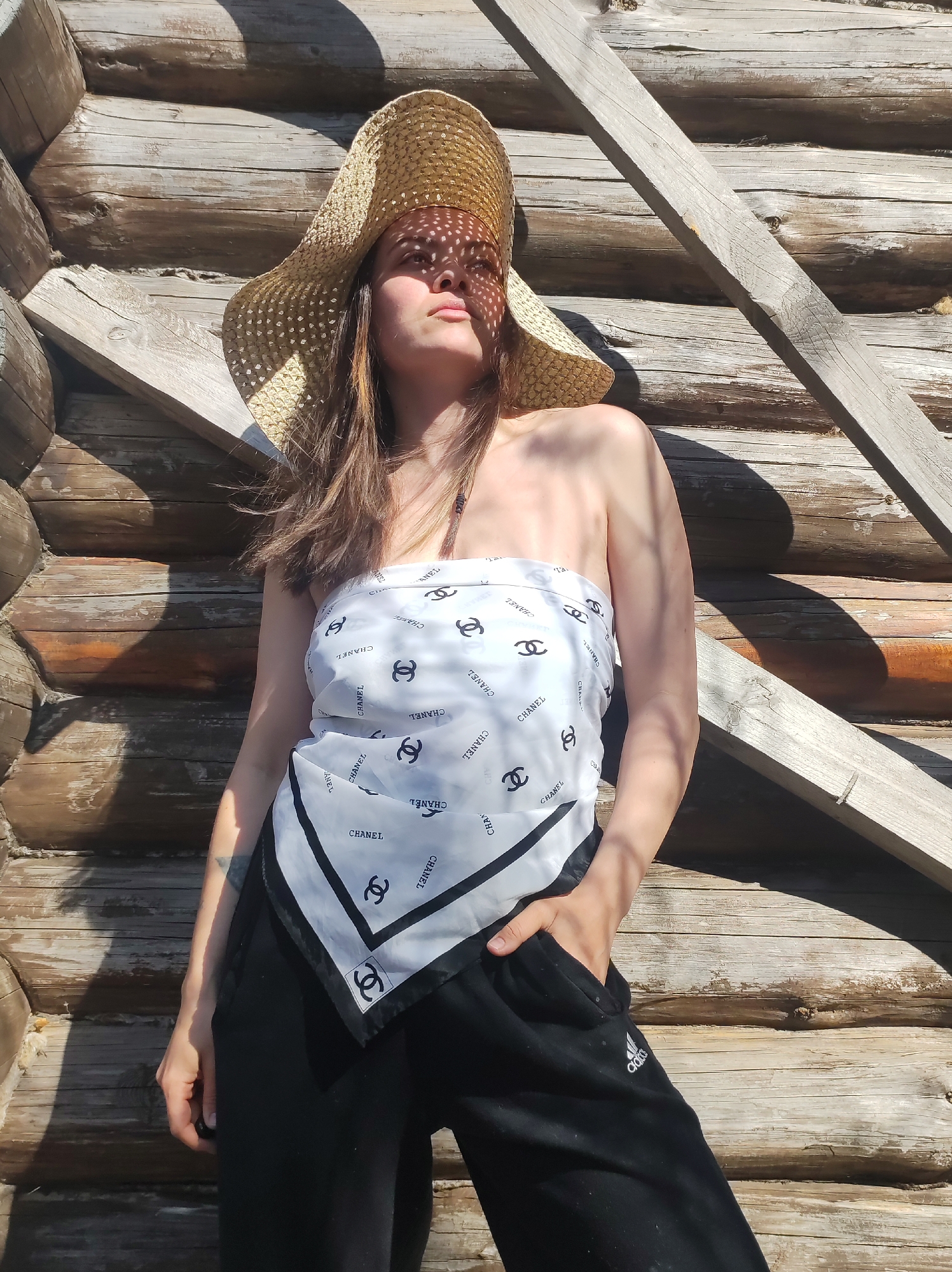 A young woman is captured in a sunlit, full-body portrait, posing against a rustic log cabin or wooden structure. She wears a wide-brimmed straw hat, which casts intricate dappled shadows across her face and upper body from the strong sunlight. Her gaze is directed upwards and slightly to the right, conveying a contemplative or serene expression.
She is dressed in a white strapless top, which appears to be a scarf tied around her chest, patterned with the "CHANEL" logo and the iconic interlocking C's. Her lower attire consists of casual black athletic pants, with the "Adidas" logo visible on her left thigh. Her right hand is casually tucked into a pocket of her pants, while her left arm hangs by her side, revealing a faint tattoo on her forearm.
The background is dominated by the textured, weathered logs of the building, accented by visible wooden beams. The bright lighting and sharp shadows suggest a clear, sunny day outdoors. The setting is identified as Фоки, Russia.