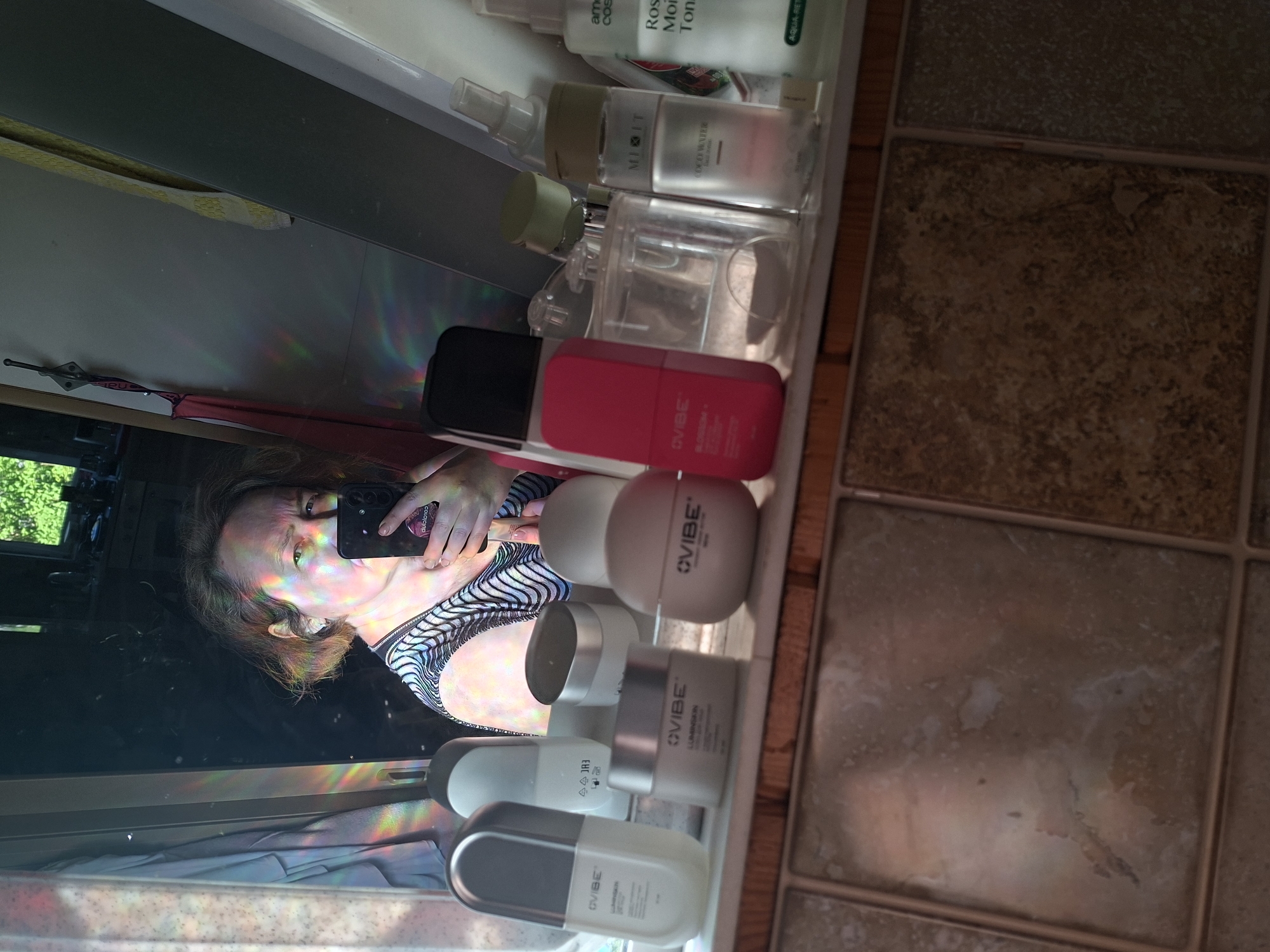 The image displays a selfie taken by a person reflected in a mirror or a similar reflective surface. A woman is visible in the reflection, holding a black smartphone with a "dulolo10" sticker on its case, capturing her own image. Her face, upper body (wearing a patterned top), and hair are illuminated by vibrant rainbow-colored light refractions that spread across the reflective surface.
A collection of cosmetic or skincare products is neatly arranged on a shelf directly in front of the reflective surface. Identifiable items include multiple "OVIBE" brand products: a pink bottle with a black cap, a round light beige container, a silver-capped container labeled "LUMIINSKIN," and a white oval container partially visible as "LUMINAIR." Other products include a clear spray bottle from "MIOIT" with "COCO WATER" printed on it, a white bottle with "Rose Mois Ton" (likely "Rose Moisture Toner"), and a bottle partially labeled "am cos" and "AQUA-RET."
To the right of the shelf, a brown and beige tiled floor is visible. The reflection also reveals green foliage from trees in the background, suggesting a daytime setting with a window overlooking an outdoor area. A yellow strap and a piece of red fabric are faintly discernible in the reflected background. The prominent rainbow light effects imply bright sunlight interacting with the environment, creating a colorful display.