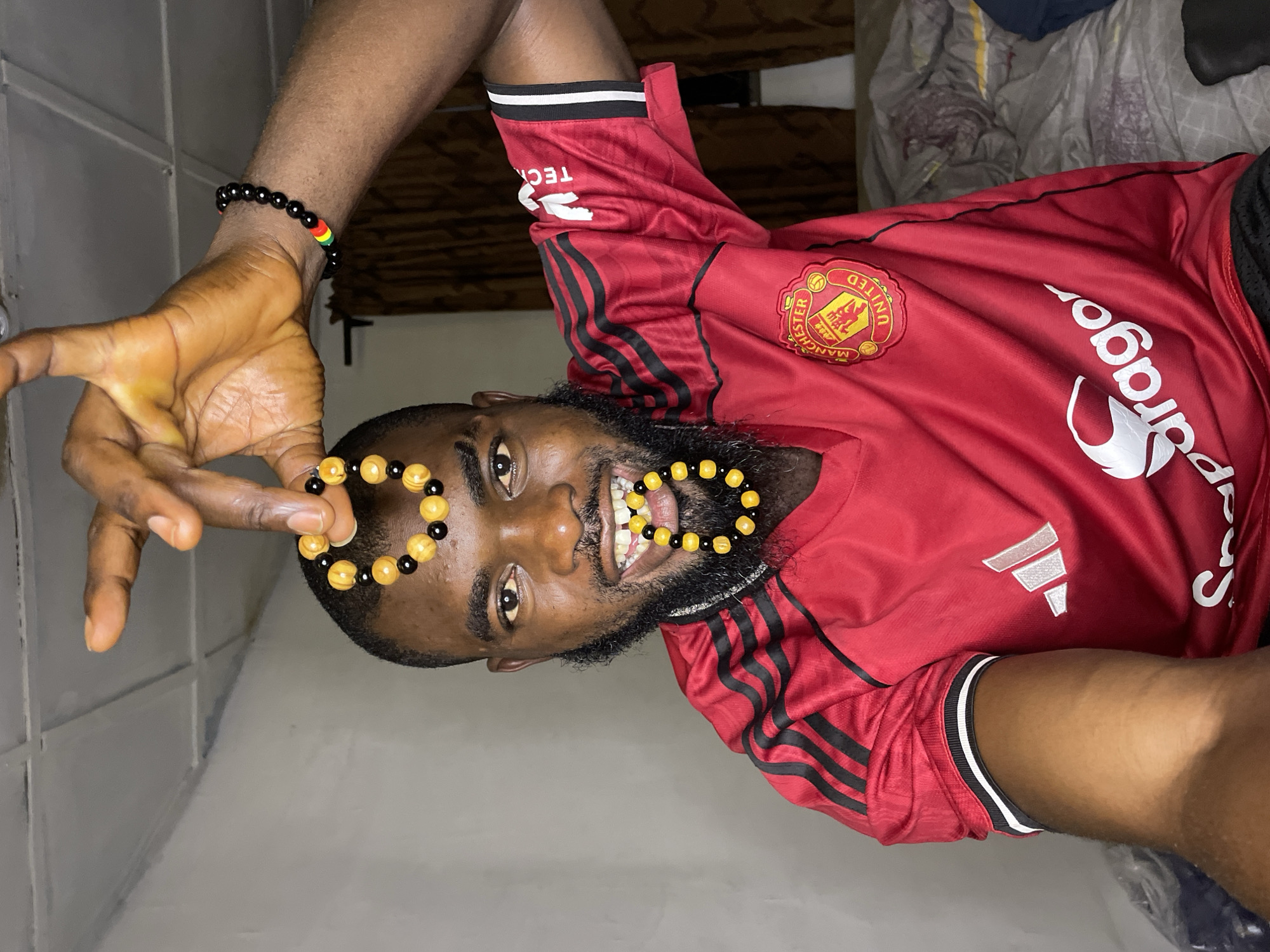 A man with dark skin and a beard is shown from the chest up, wearing a red Manchester United jersey. He is holding up two bracelets made of yellow and black beads, one above his head and another across his mouth. He has a black bracelet with red, yellow, and green beads on his left wrist. The background appears to be an indoor setting, possibly a bedroom, with a textured ceiling and a window with curtains visible. The lighting is dim. The image's context does not provide any information about the city of Jos, Nigeria.