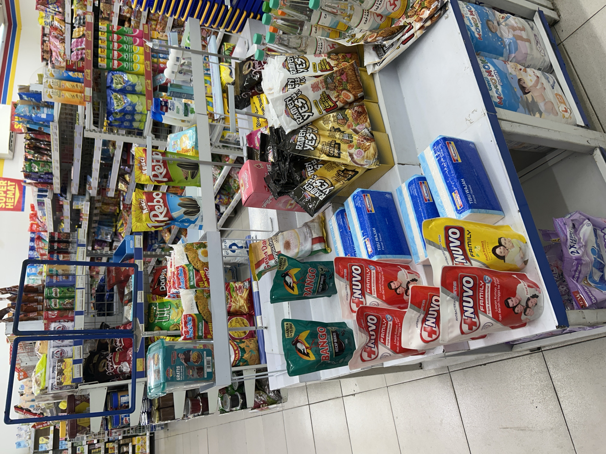 The image shows a wide shot of a retail store, likely an Indomaret convenience store, given the branding visible on some products. The store is brightly lit, with rows of shelves filled with packaged food items, drinks, and toiletries. The floor is tiled with off-white and grey squares.
In the foreground, on a tiered display rack, are several products. On the top shelf, there are two green pouches of Bango Kecap Manis (sweet soy sauce) and a pack of four blue and white packaged facial tissues. To the right of the tissues are two red pouches of Nuvo Family Triple Antibacterial body wash, and to the right of those is a yellow pouch of Nuvo Family Fresh Protect body wash. Below these, on a lower shelf, are additional packages of facial tissues and diapers.
On the shelves behind the foreground display, a variety of packaged snacks and instant noodles are visible, including brands like Indomie, Rebo, and Doritos. There are also bottles of drinks on the shelves. The store appears well-stocked. There are no people visible in the image. The overall impression is a clean and organized retail environment. The presence of multiple Indomaret branded items strongly suggests the location is within one of their stores in Depok, Indonesia.