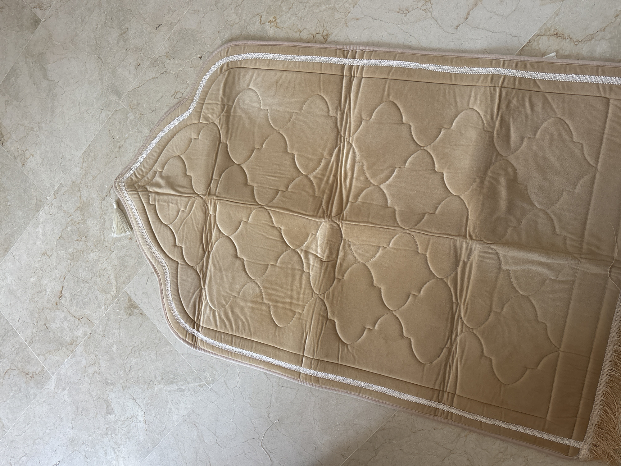 A beige prayer rug with a white decorative trim and tassels lies on a marble-tiled floor. The rug has a quilted pattern of quatrefoil shapes. The floor tiles are large and rectangular, laid in a diagonal pattern, with subtle variations in color and veining characteristic of marble. The lighting suggests an indoor environment, possibly a home or a place of worship. There are no people visible in the image. The location of Orihuela, Spain, is not discernible from the visual information provided in the image.