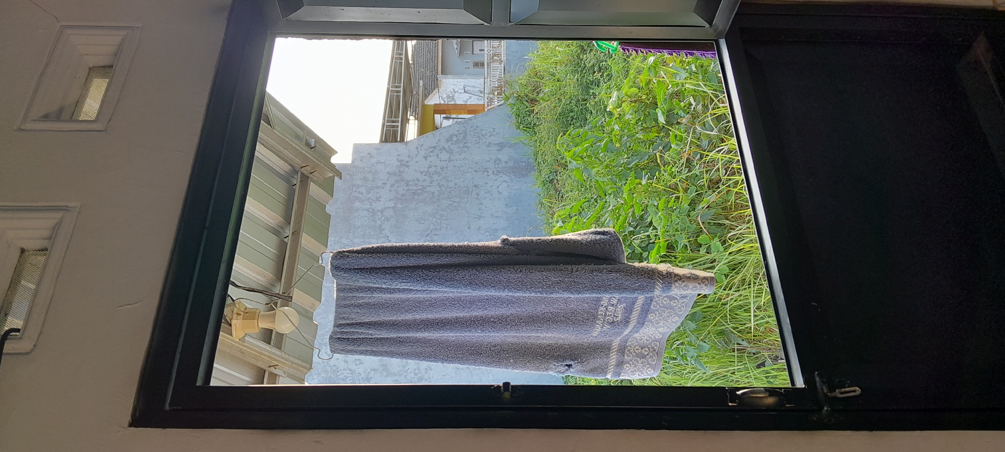The image shows a view through an open window or doorway looking out onto an outdoor area. In the foreground, a gray towel is hanging on a metal hanger, suspended from a light fixture attached to a corrugated metal roof. The towel has a patterned border at the bottom. The background consists of a lush green overgrown area with tall grass and plants. Beyond the vegetation, there are buildings with concrete walls and pitched roofs, suggesting a residential or semi-urban environment. The lighting indicates it is daytime, with sunlight casting shadows. The location is identified as Tambakrejo, Indonesia. The scene is a candid snapshot of everyday life, possibly depicting laundry drying.