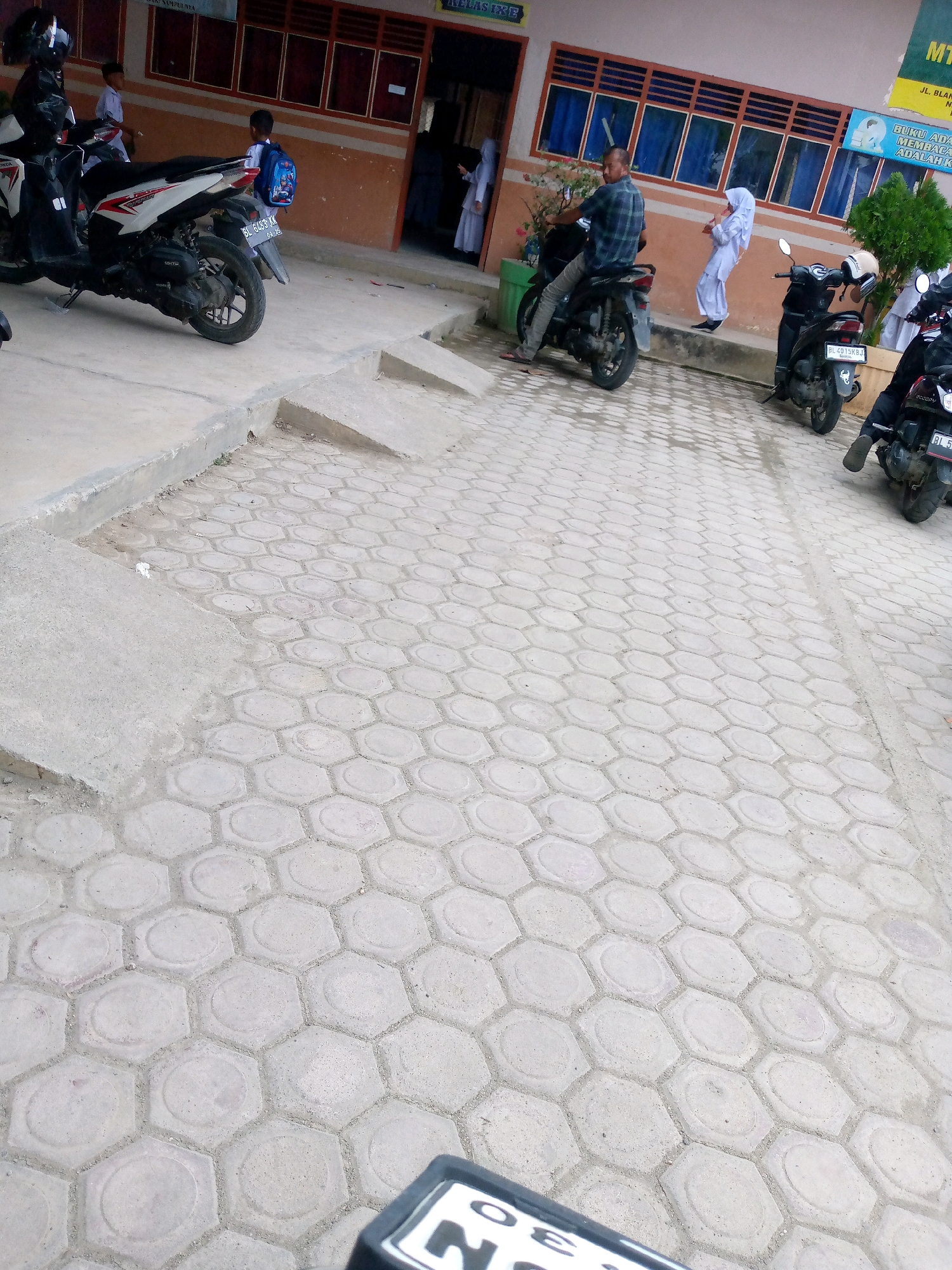 The image captures an outdoor scene, likely a school courtyard in Mesjid, Indonesia, paved with hexagonal concrete tiles. In the foreground, part of a motorcycle's license plate is visible, displaying "30". Several parked motorcycles are present. On the left, a white and red scooter is parked, with a black helmet on its handlebars. Further back, a man in a checkered shirt rides a motorcycle, while a person in white religious attire stands near a potted plant. Another motorcycle is parked with a white helmet and a cream-colored seat. In the background, a building with brown walls and rows of blue-paned windows is visible. A sign in yellow and green is attached to the building, with text in Indonesian. Children in school uniforms are also seen in the vicinity. The overall atmosphere suggests a typical day at a school with students and visitors arriving or departing on motorcycles.