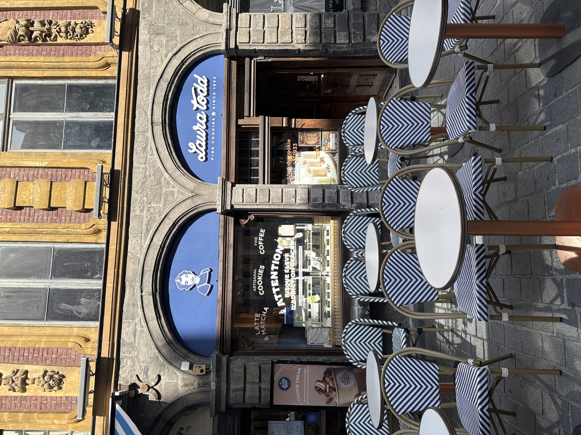 The image shows an outdoor seating area in front of a bakery called "Laura Todd" on a cobblestone street in Lille, France. The bakery has a blue facade with the bakery's name and logo, and large display windows showcasing baked goods. Several small, round tables with white tops and copper-colored legs are set up, each surrounded by chairs with woven blue and white chevron patterned seats and backs. The chairs are made of wicker with light-colored frames. The architecture of the building is historic, with ornate yellow stone detailing above the windows and doors. The lighting suggests it is daytime, with strong shadows cast on the ground. No people are visible in the scene, but the presence of tables and chairs implies it is a public establishment.
