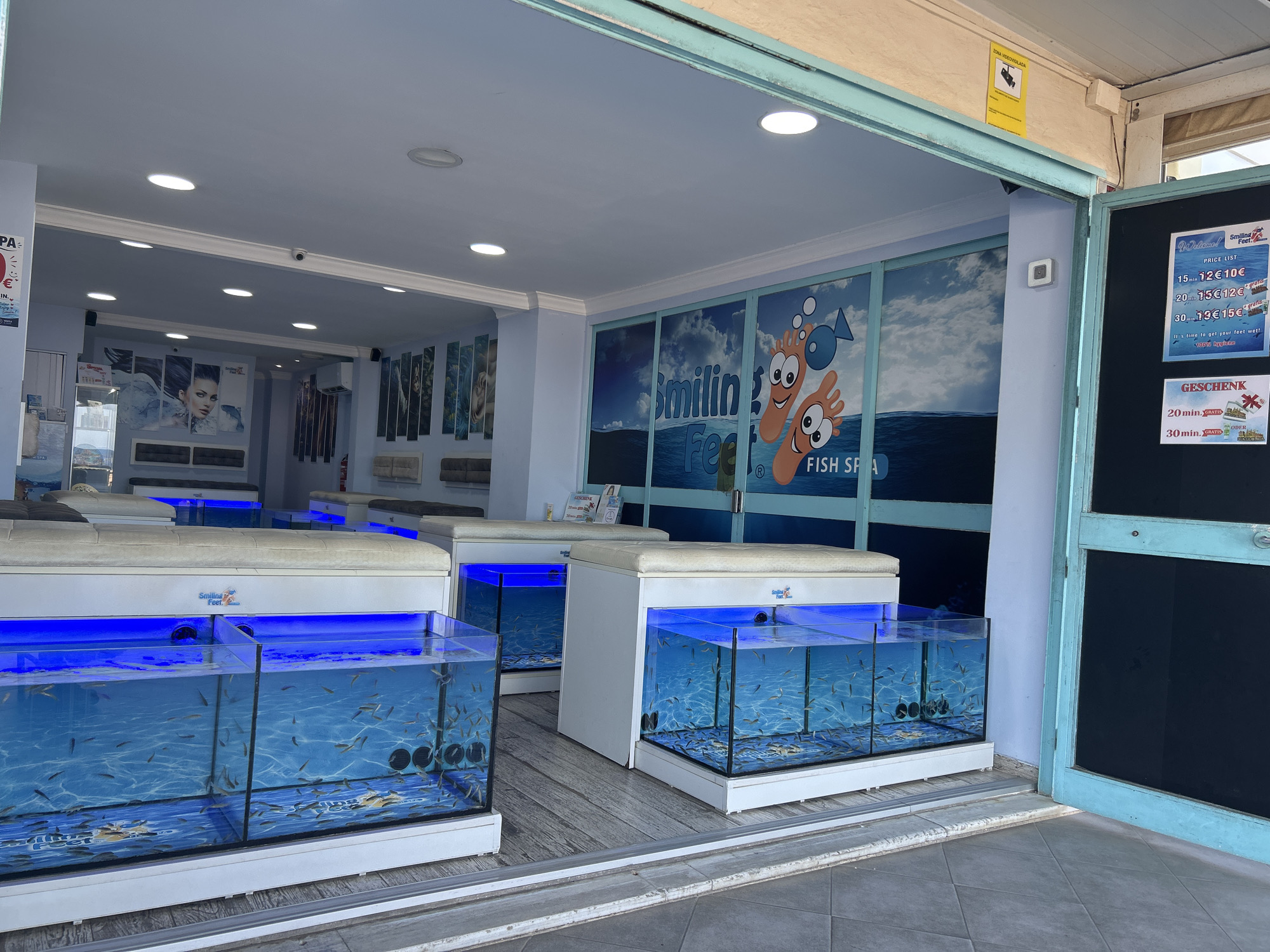The image shows the exterior of a fish spa establishment named "Smiling Feet Fish Spa" located in Cala Rajada, Spain. The storefront features large glass windows displaying multiple fish tanks filled with small fish and illuminated by blue LED lights. These tanks are positioned in front of white benches where customers would sit for the fish pedicure service. A price list board is visible on the right side of the entrance, detailing prices for different durations of the service. The interior of the spa is decorated with images of models and aquatic themes. The surrounding environment appears to be an outdoor walkway or street.