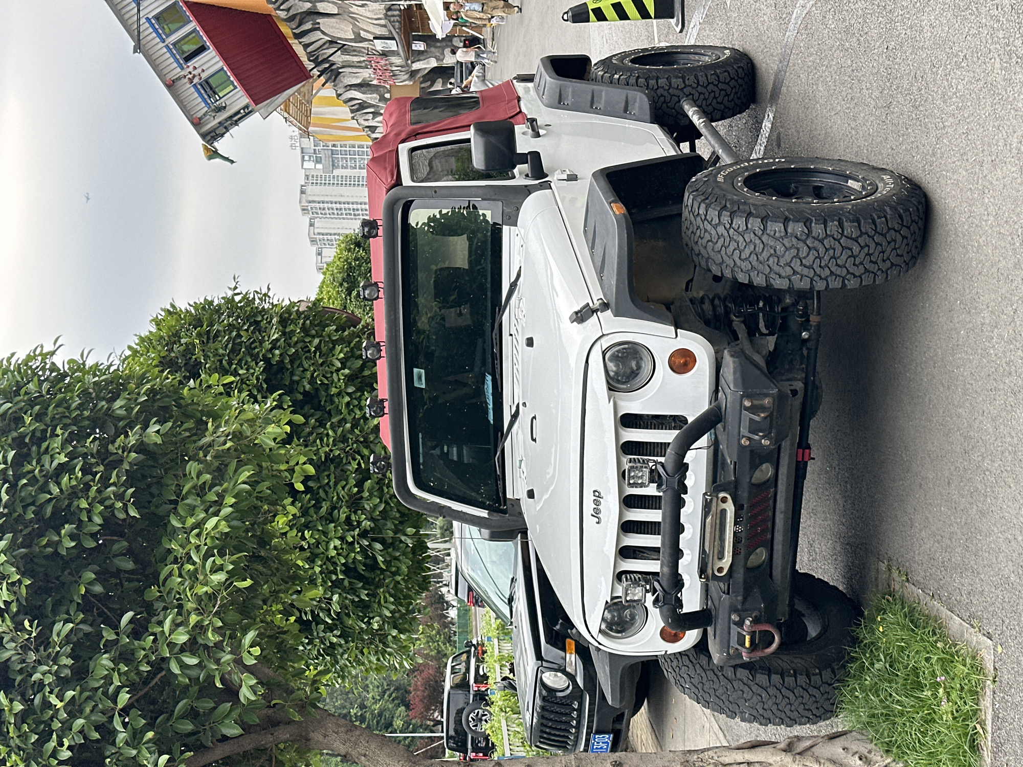 A white Jeep with a red canvas top is parked on a city street in Nan'an District, China. The vehicle is parked on the right side of the road, next to a curb with green grass. The front of the Jeep faces the viewer, showcasing its black bumper, winch, and distinctive grille. A black Jeep is parked behind the white Jeep. A tree with dense green foliage partially obscures the view on the left side. Across the street, there are buildings with unusual architectural features and vibrant colors. Tall buildings can be seen in the background. The sky is overcast.