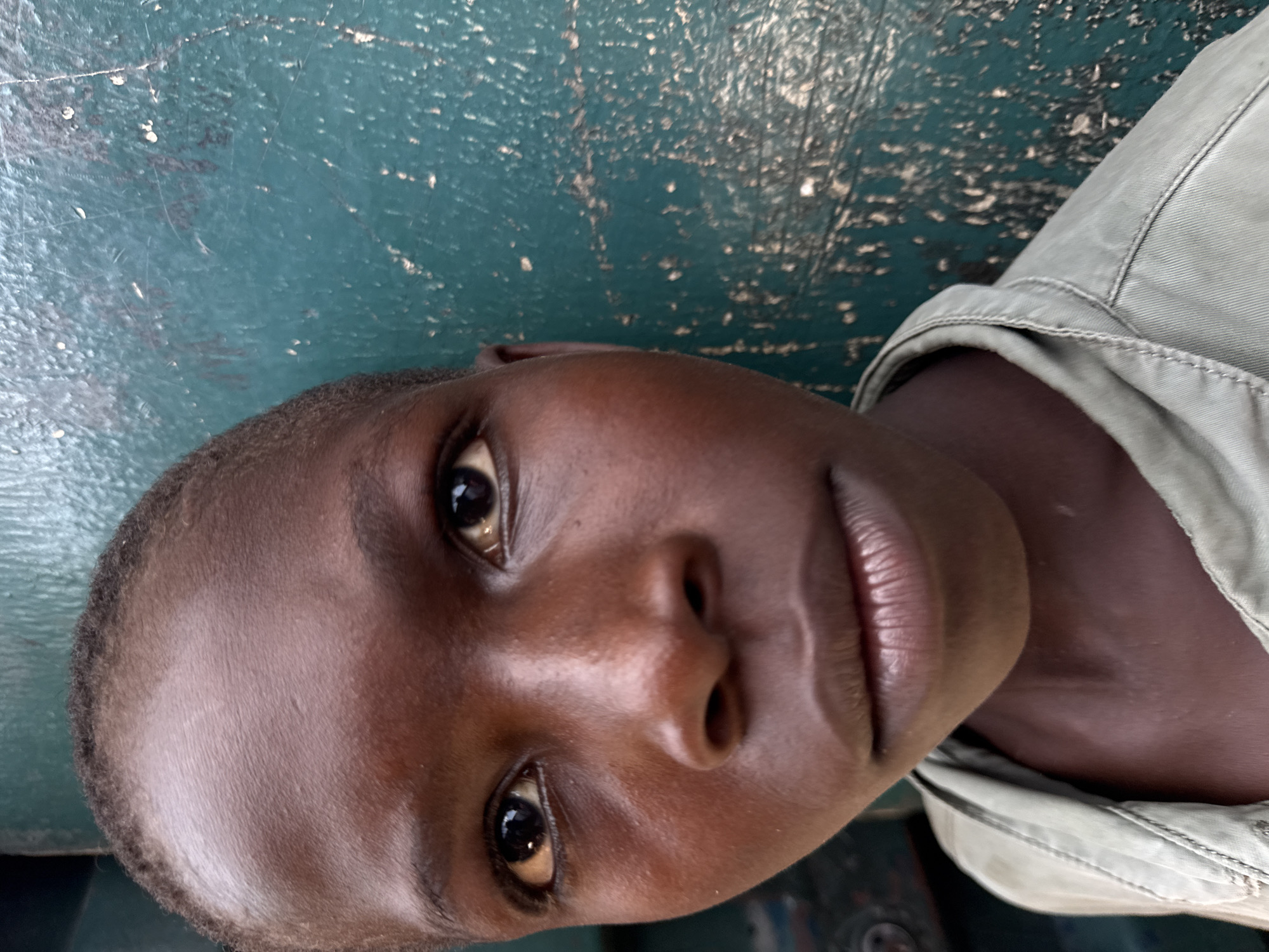 A close-up, rotated portrait of a young, dark-skinned individual with dark eyes and short hair. The individual is wearing a light-colored shirt. The background appears to be a textured green surface. The location and city of Lekki, Nigeria, cannot be confirmed or denied from the image alone.
