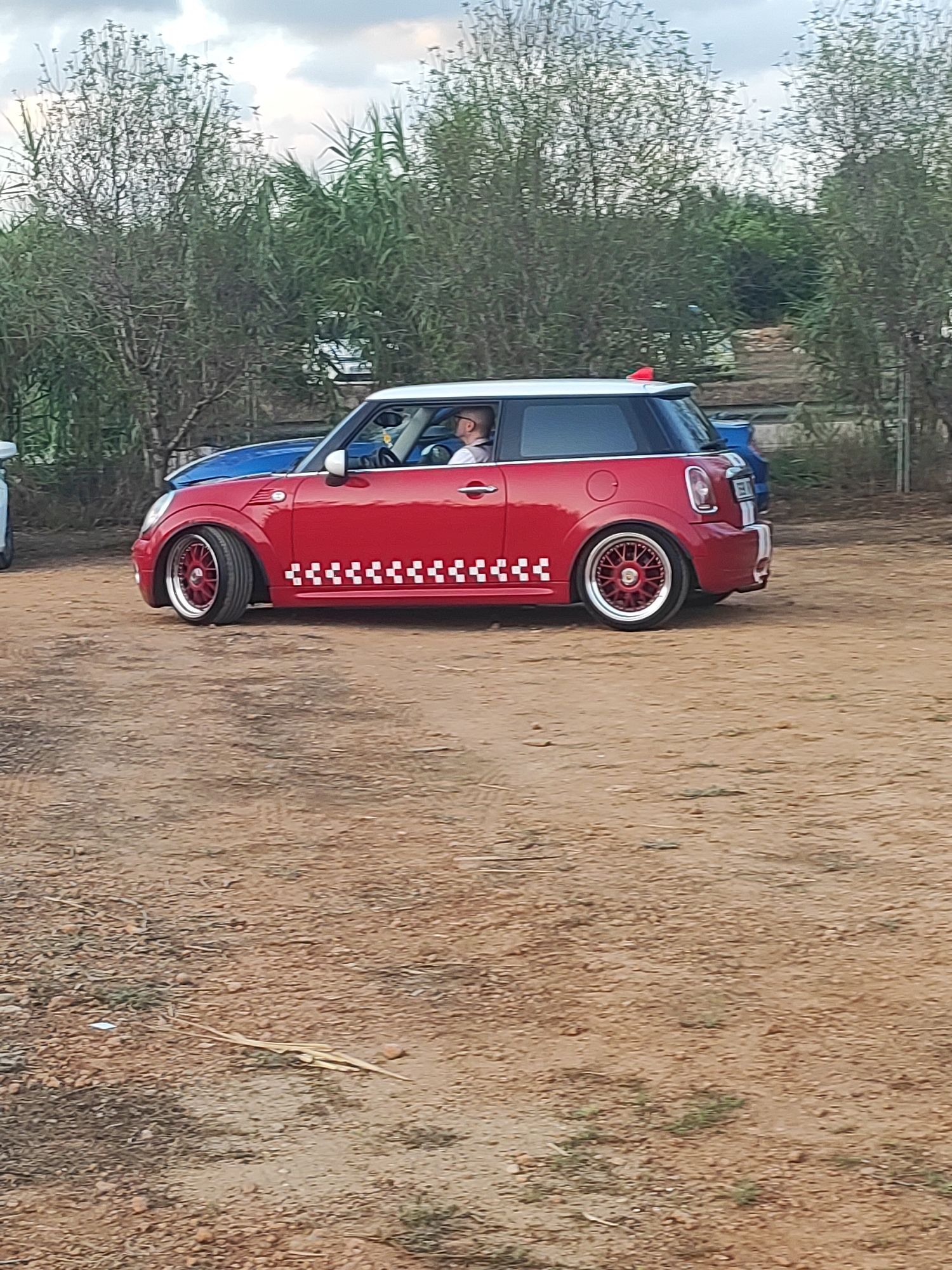 A red Mini Cooper, seen from the side, is parked on a dirt ground, likely in a rural or undeveloped area near Alcàsser, Spain. The car features a white roof and racing stripes on the rear. A person is visible inside the car, in the driver's seat, wearing glasses. The car's wheels are custom red alloy rims. In the background, there are trees and vegetation, and a blue car is partially visible behind the Mini Cooper. The sky is cloudy.