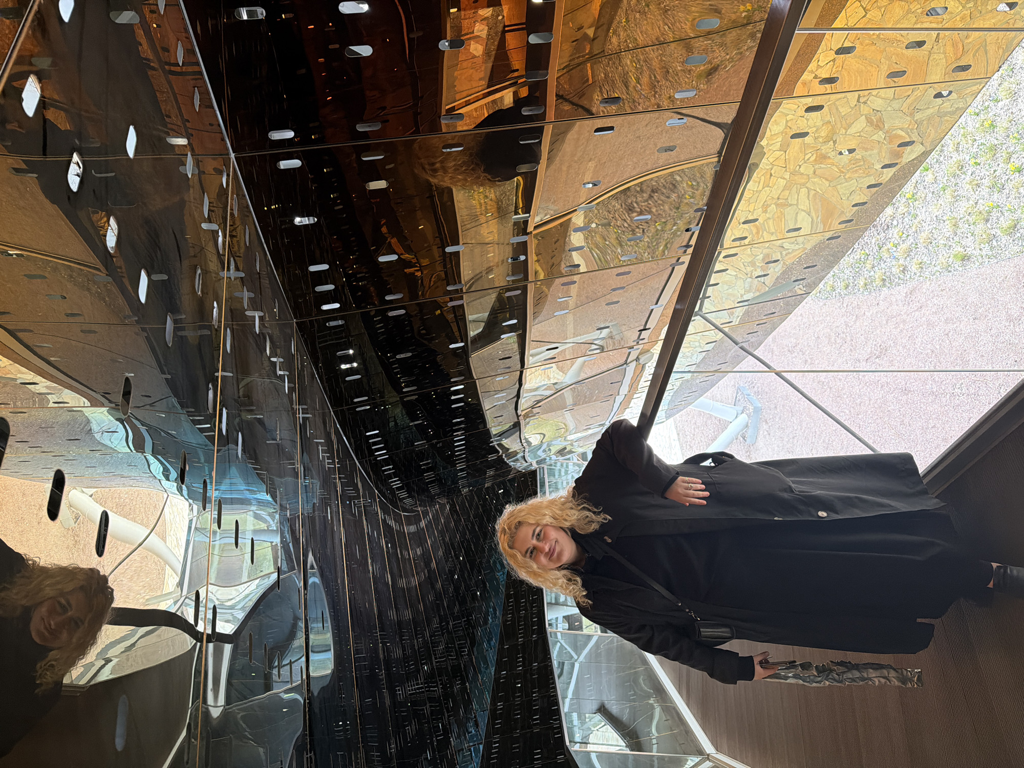 The image depicts a woman with curly blonde hair, dressed in black attire, standing in a reflective space. The reflective surfaces create distorted reflections of the surrounding environment, including exterior elements and the woman herself. The scene gives the impression of an art installation or a contemporary architectural design. The location is the Zaryadye Park in Moscow, Russia.