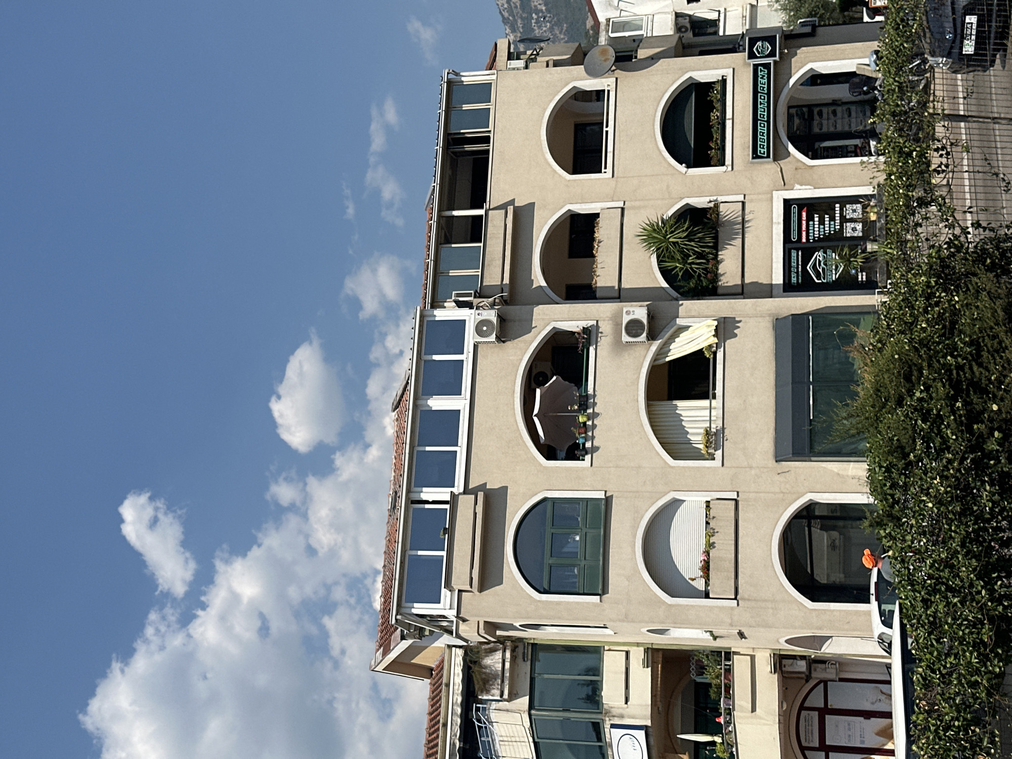 In Budva, Montenegro, a beige building with multiple arched windows and balconies is depicted under a clear blue sky with scattered white clouds. The building appears to be a commercial and residential structure, with storefronts at the ground level. One storefront is labeled "CABRIO AUTO RENT," and another displays "RENT A CAR". Above the ground floor, several windows and balconies are visible, some adorned with plants and an umbrella. An air conditioning unit is mounted on the building's facade. In the foreground, dense green foliage obscures the lower portion of the building, and a black car is partially visible to the right. A person wearing an orange hat is seen behind the foliage, near a white car. The building's architecture features a light beige exterior, terracotta roof tiles, and white trim around the windows. The overall scene suggests a sunny day in a coastal urban environment.
