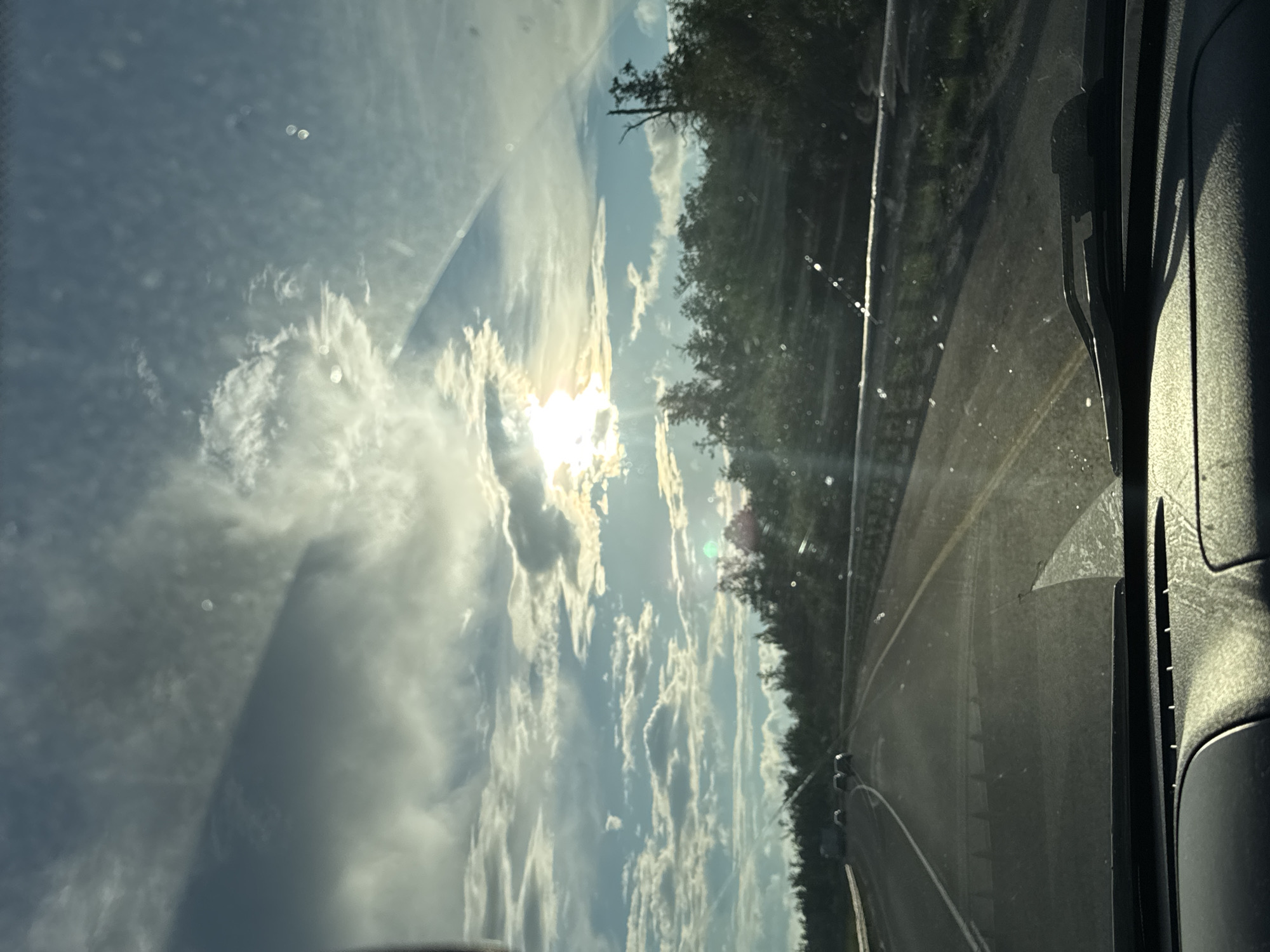 The image depicts a view from inside a vehicle driving on a highway. The sun is setting, casting a warm glow through the clouds and glinting off the windshield, which has several water spots. The highway curves to the left, with trees lining the right side of the road. Several vehicles are visible on the road ahead. The dashboard of the car is visible in the foreground. The scene appears to be in Krasnokamsk, Russia, given the context provided.